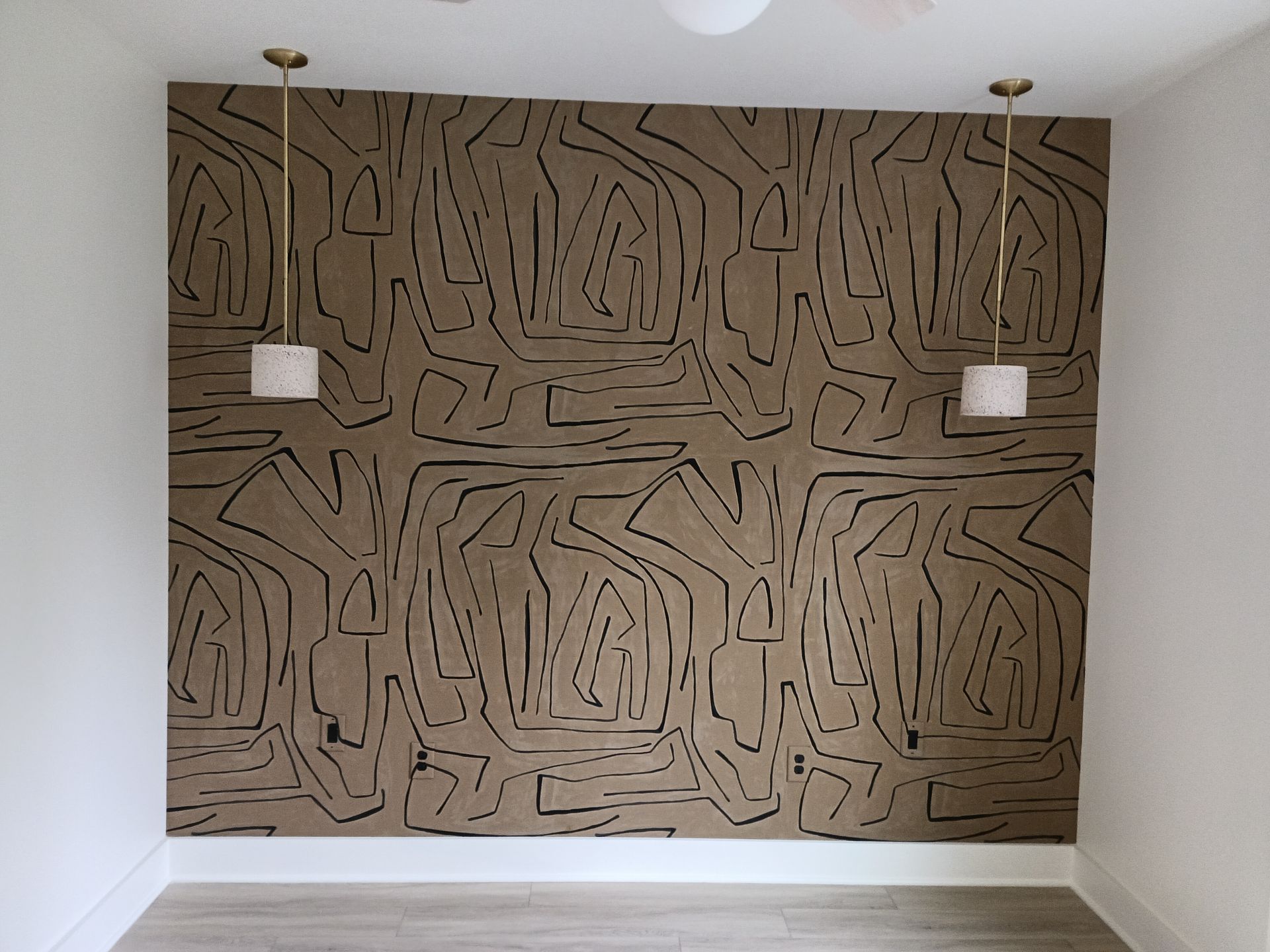 A large tan wall hanging with black lines is centered between two pendant lights in a white room.