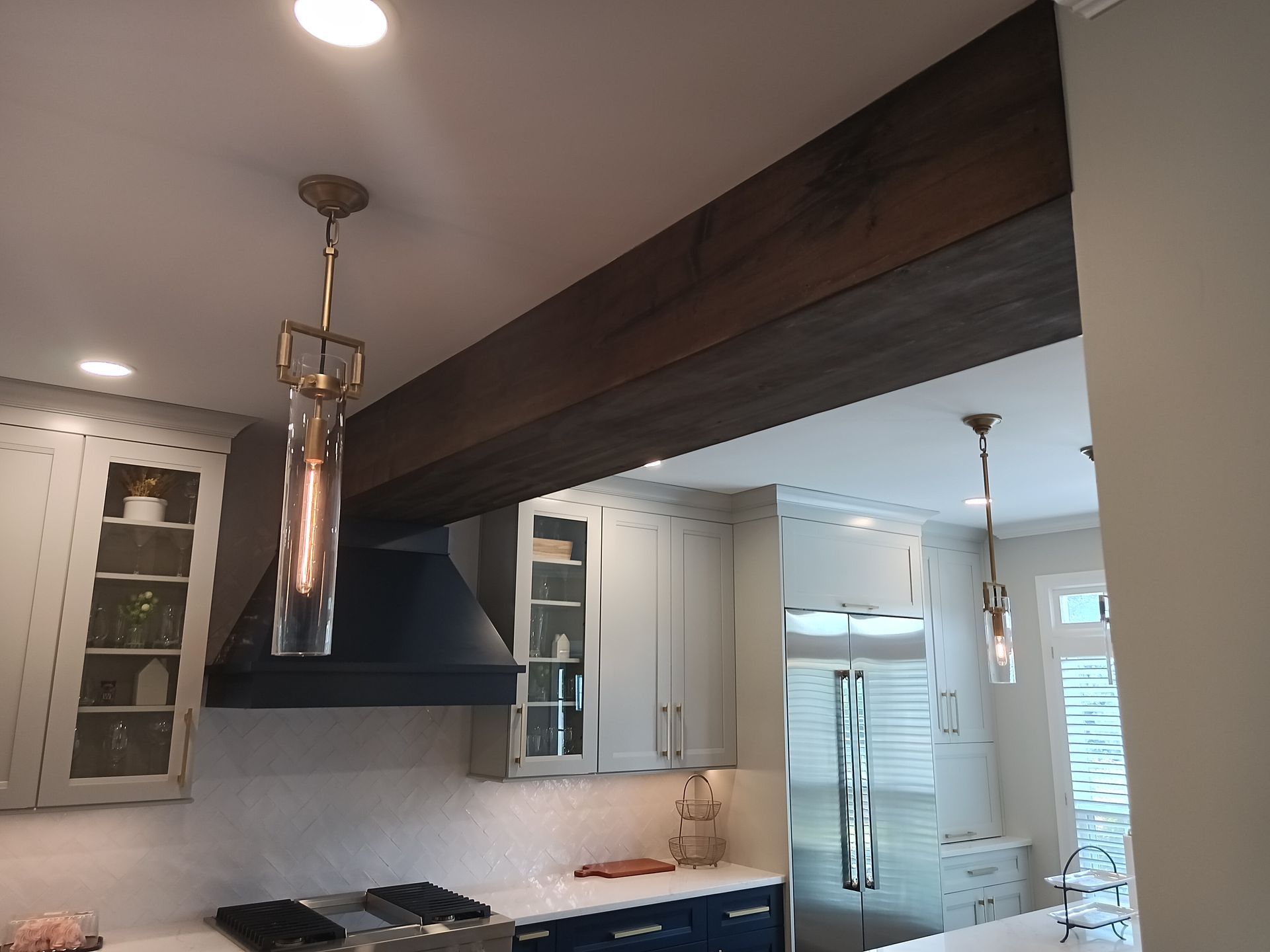 Kitchen with dark wood beam, light blue cabinets, black range hood, pendant lights, and stainless steel refrigerator.