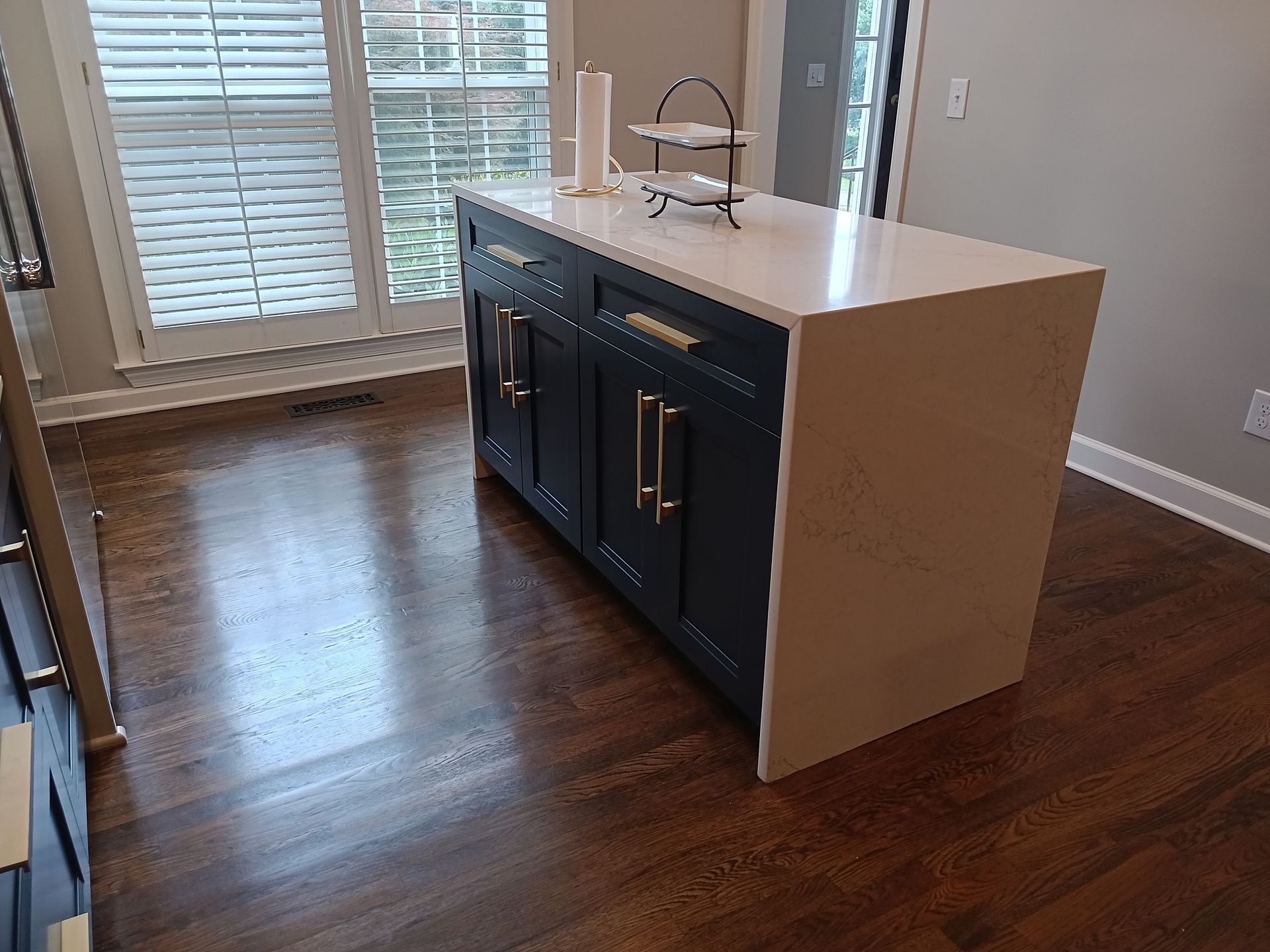 Kitchen island with navy blue cabinets, gold handles, and white countertop. Dark hardwood floor.