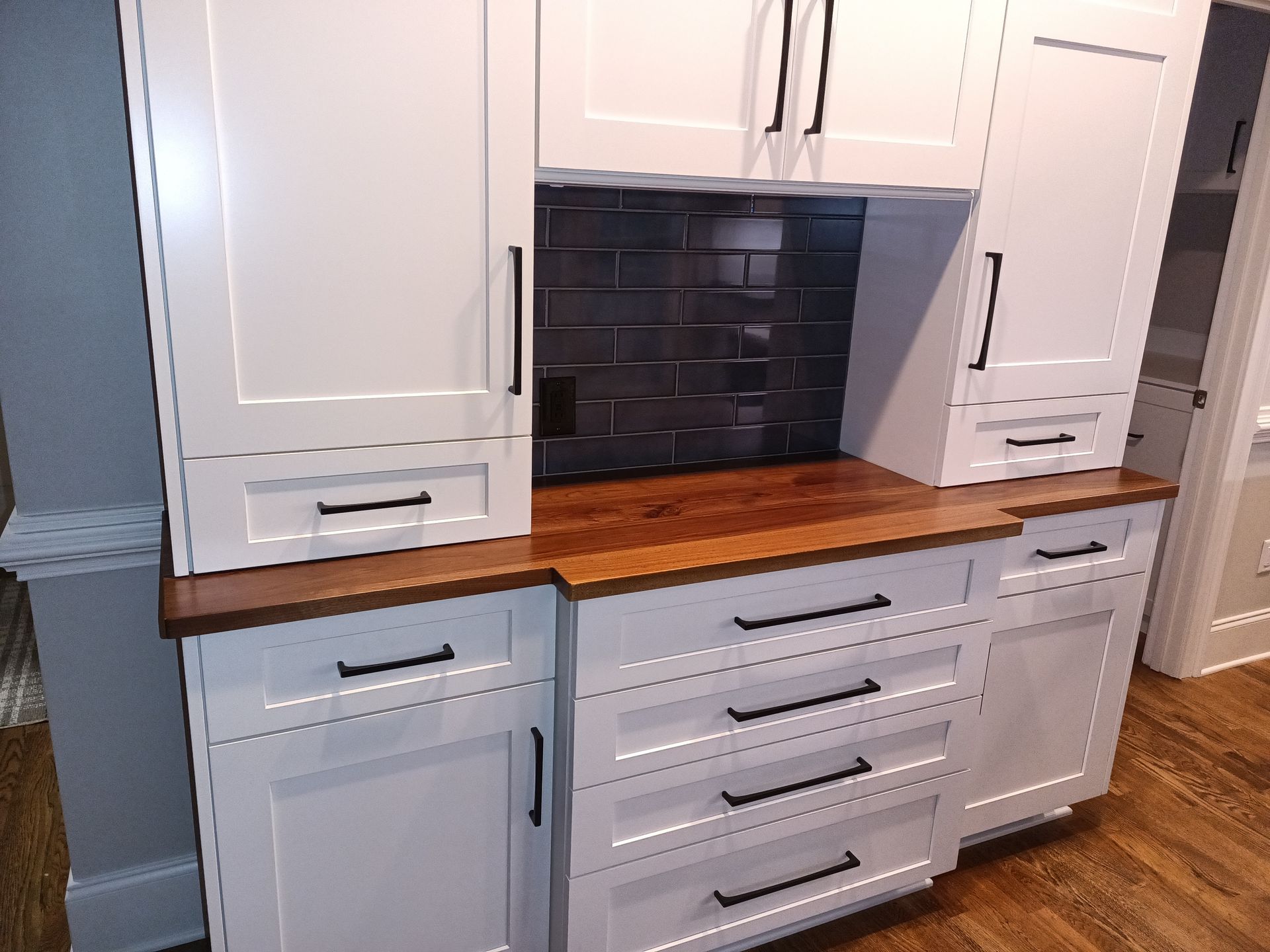 White kitchen cabinets with black hardware, wood countertop, and dark tiled backsplash.