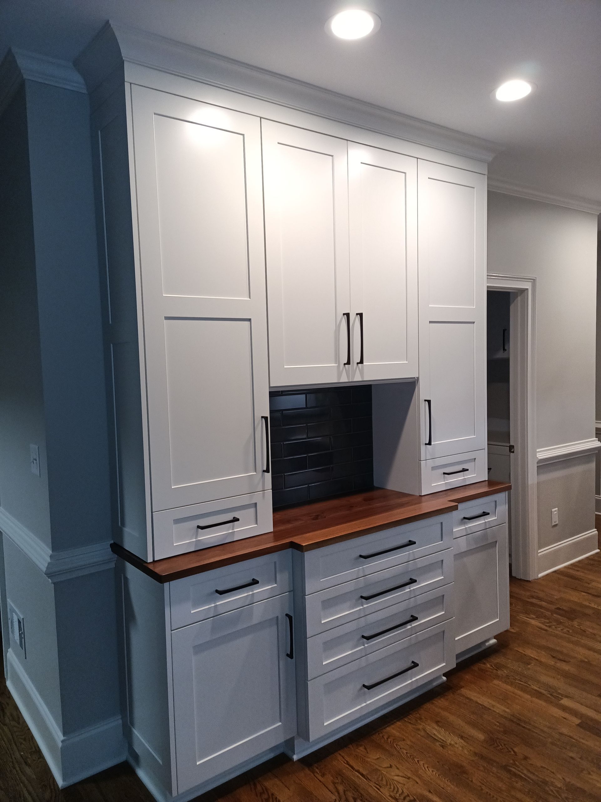 White built-in cabinets with a wooden countertop. Black hardware and backsplash, set against a neutral wall.