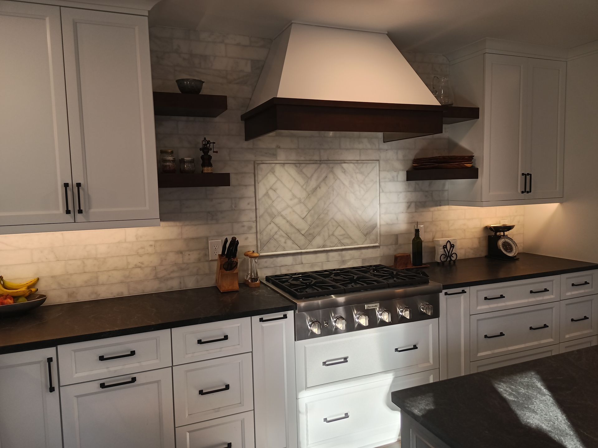 White kitchen with black countertops, gas range, wood range hood, and white cabinets.