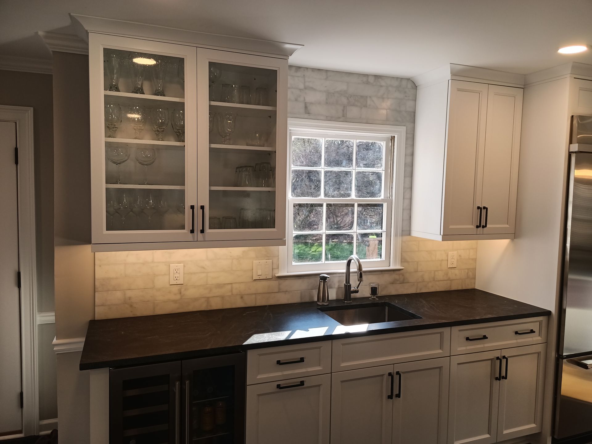 Kitchen with white cabinets, dark countertop, sink, and wine fridge. A window is above the sink, with brick backsplash.