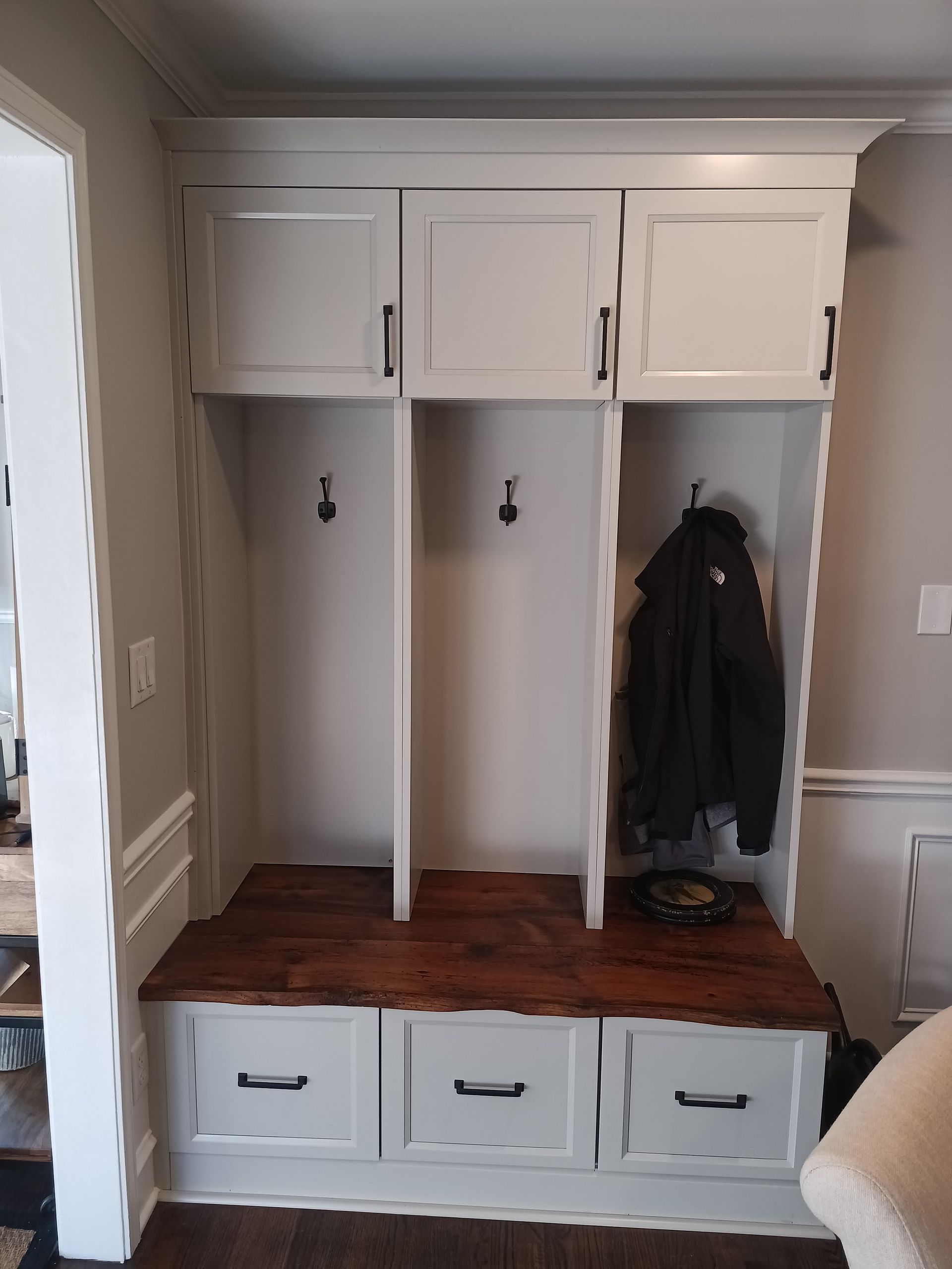 Built-in white mudroom cabinet with dark wood bench seat and three drawers below. Three compartments with hooks and overhead storage.