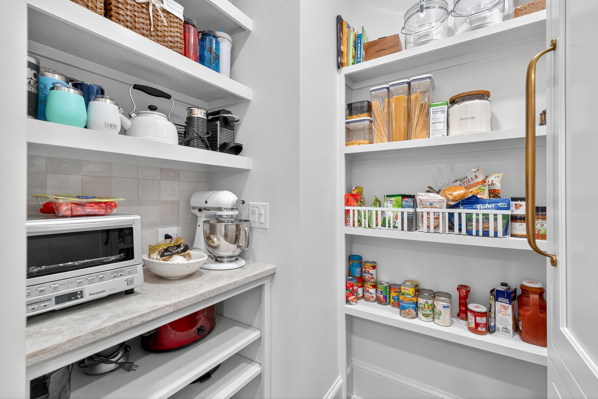 Pantry with white shelves filled with food items, appliances, and storage containers. Light gray walls.