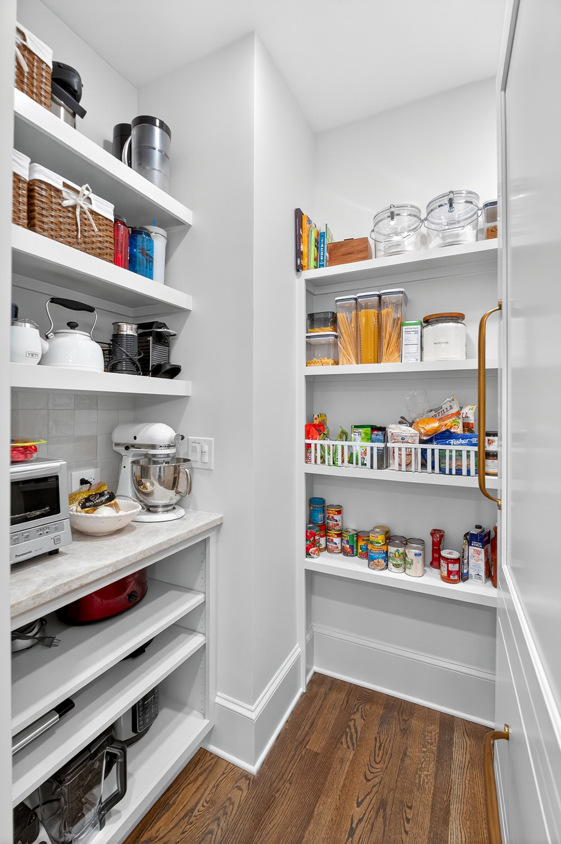 A pantry with white shelves filled with food and appliances; wood floor, white walls.