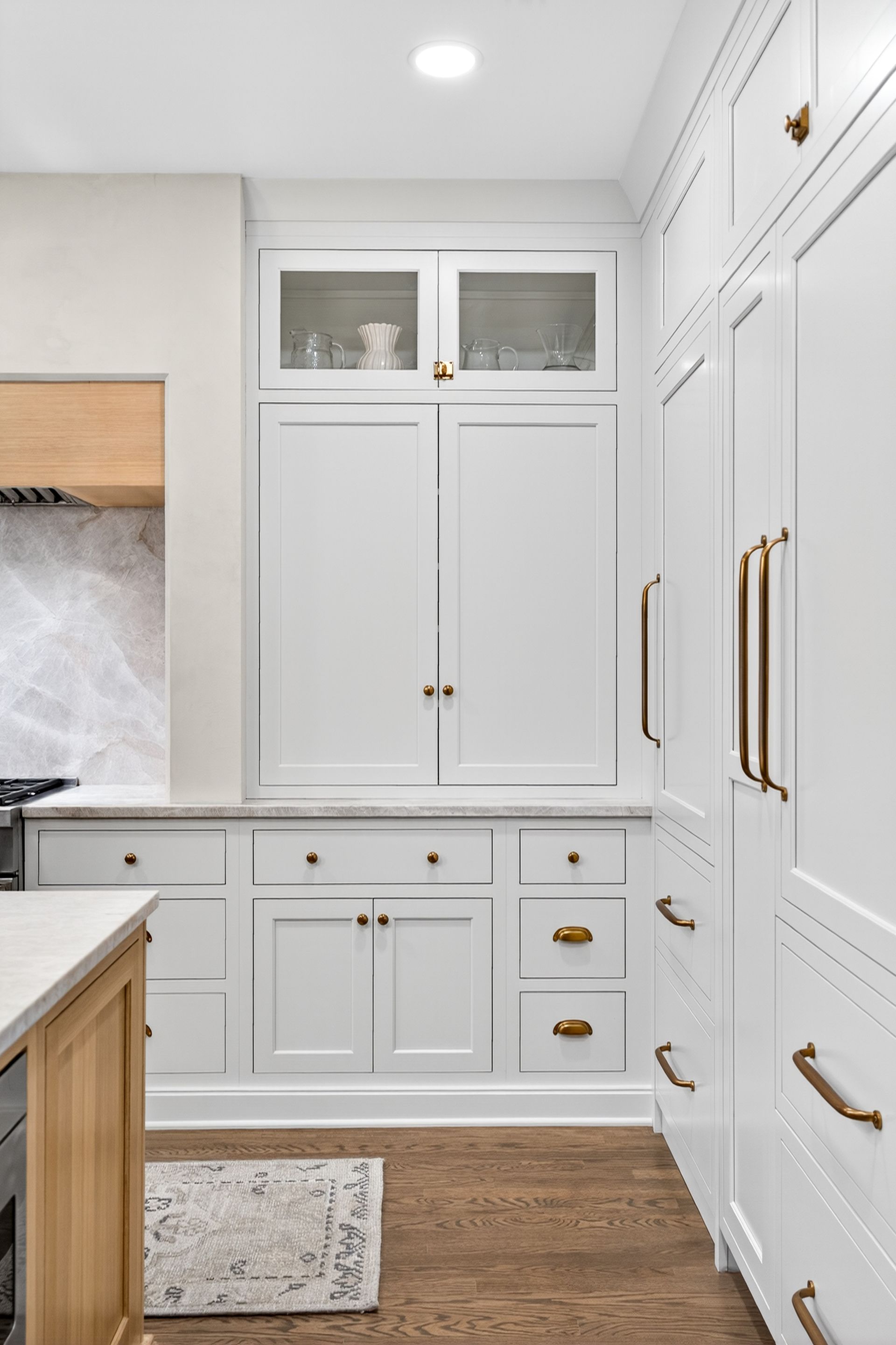 White kitchen cabinets with gold hardware and glass-front upper cabinet, over hardwood floor.