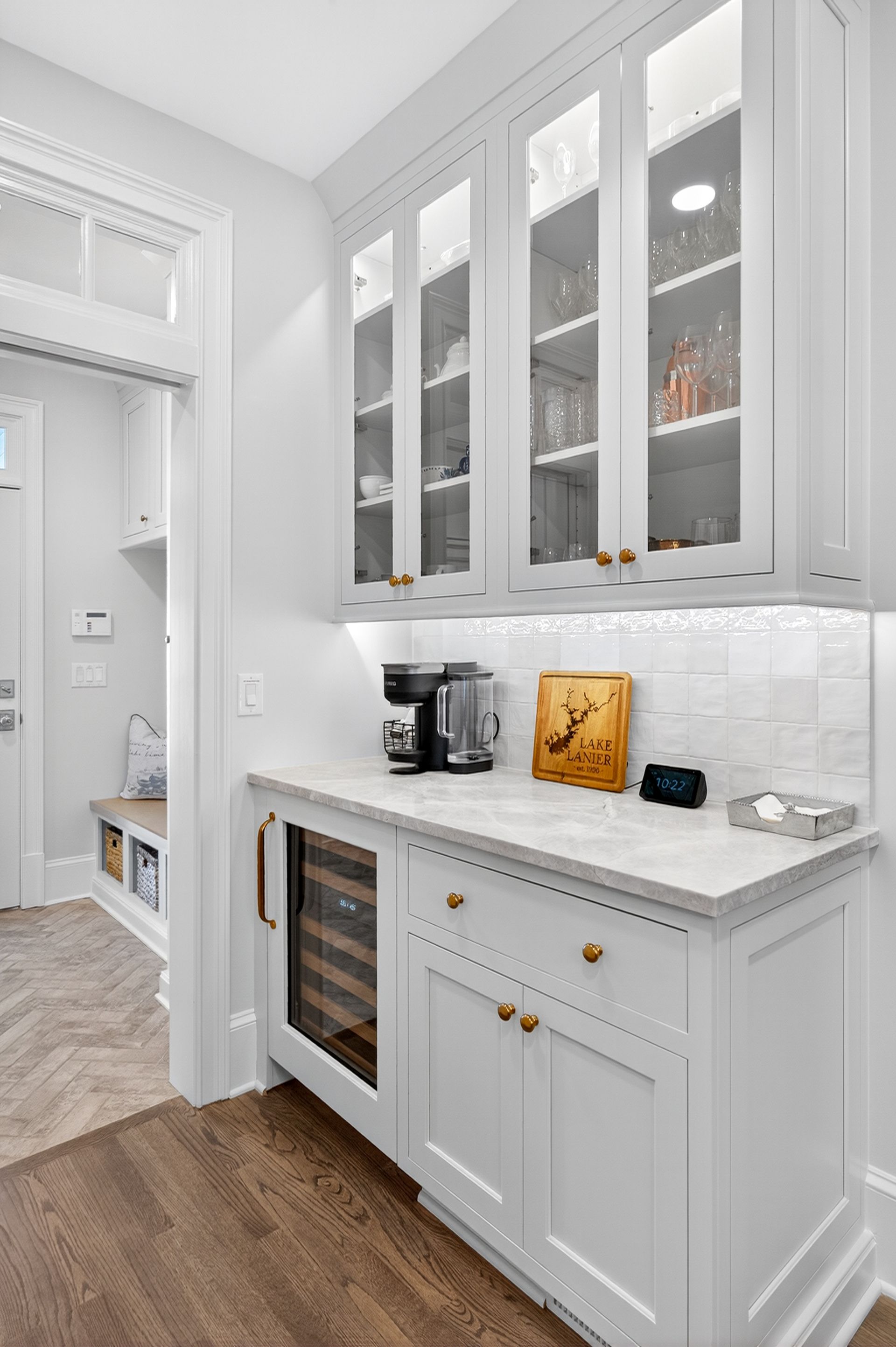 White pantry with glass-front cabinets, countertops, and a wine fridge. Includes coffee maker and decor.