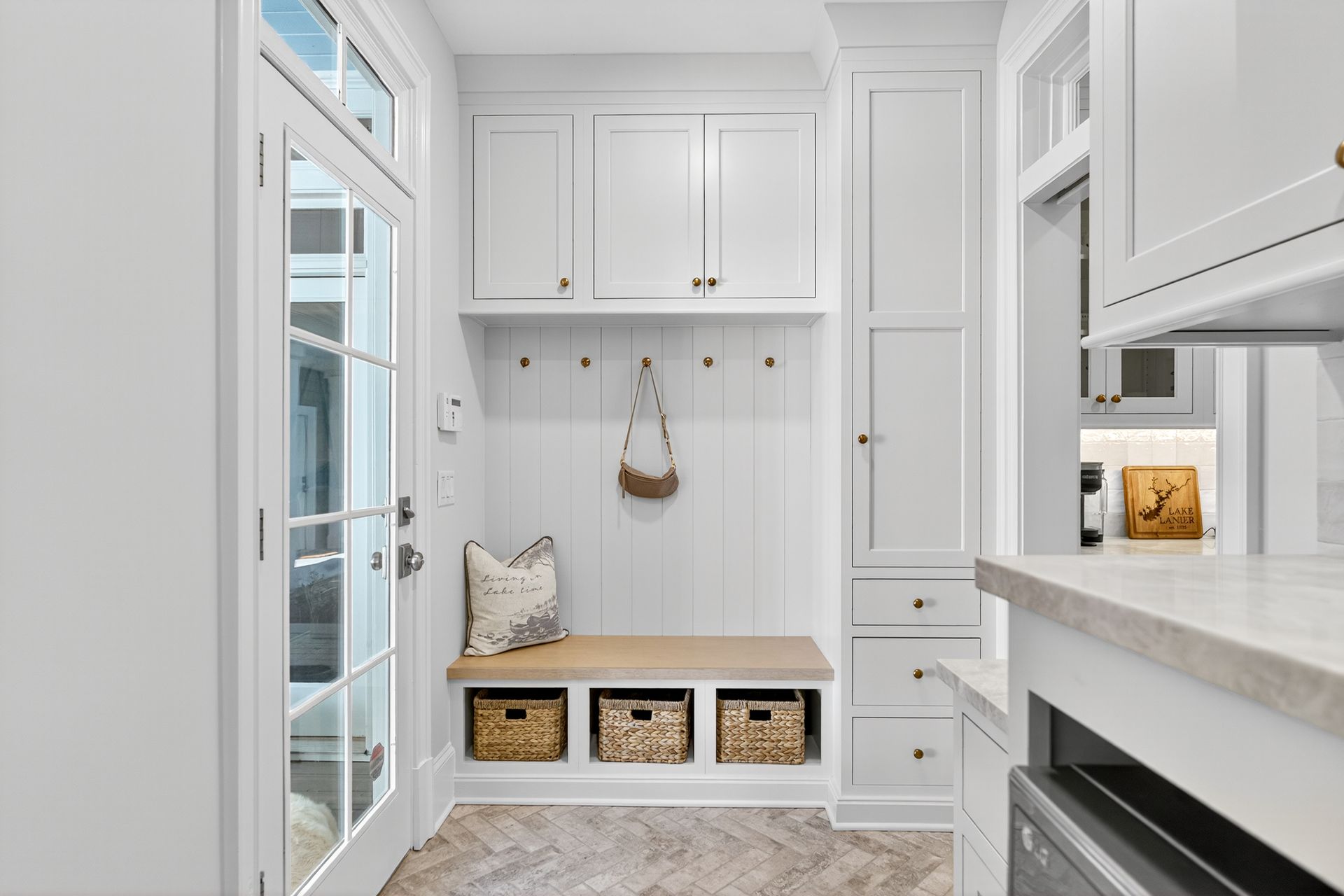 White mudroom with bench, storage, and hooks; a door to the left and cabinets to the right.