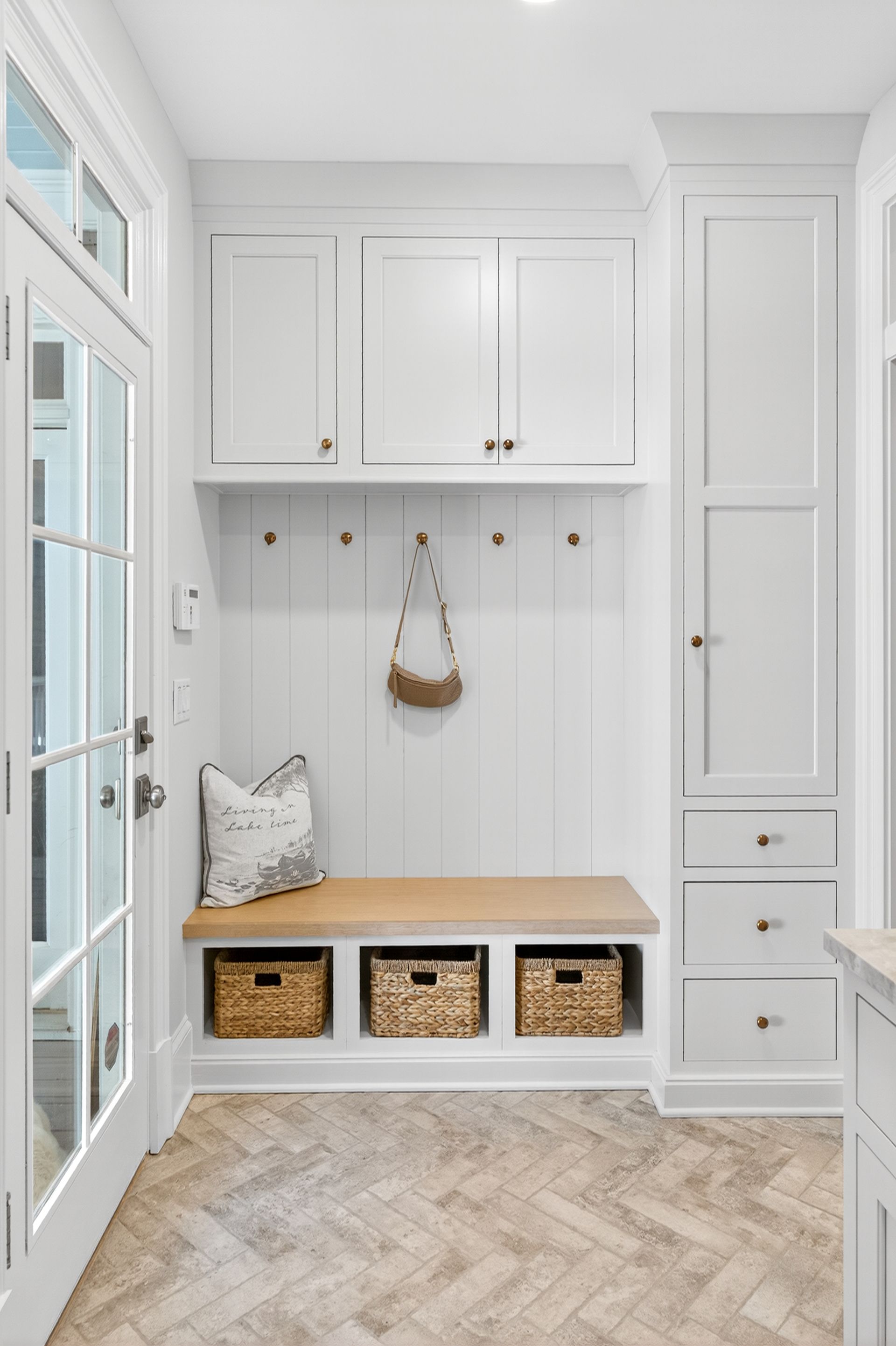 White entryway with built-in bench, storage, and herringbone floor. A bag hangs on a hook.