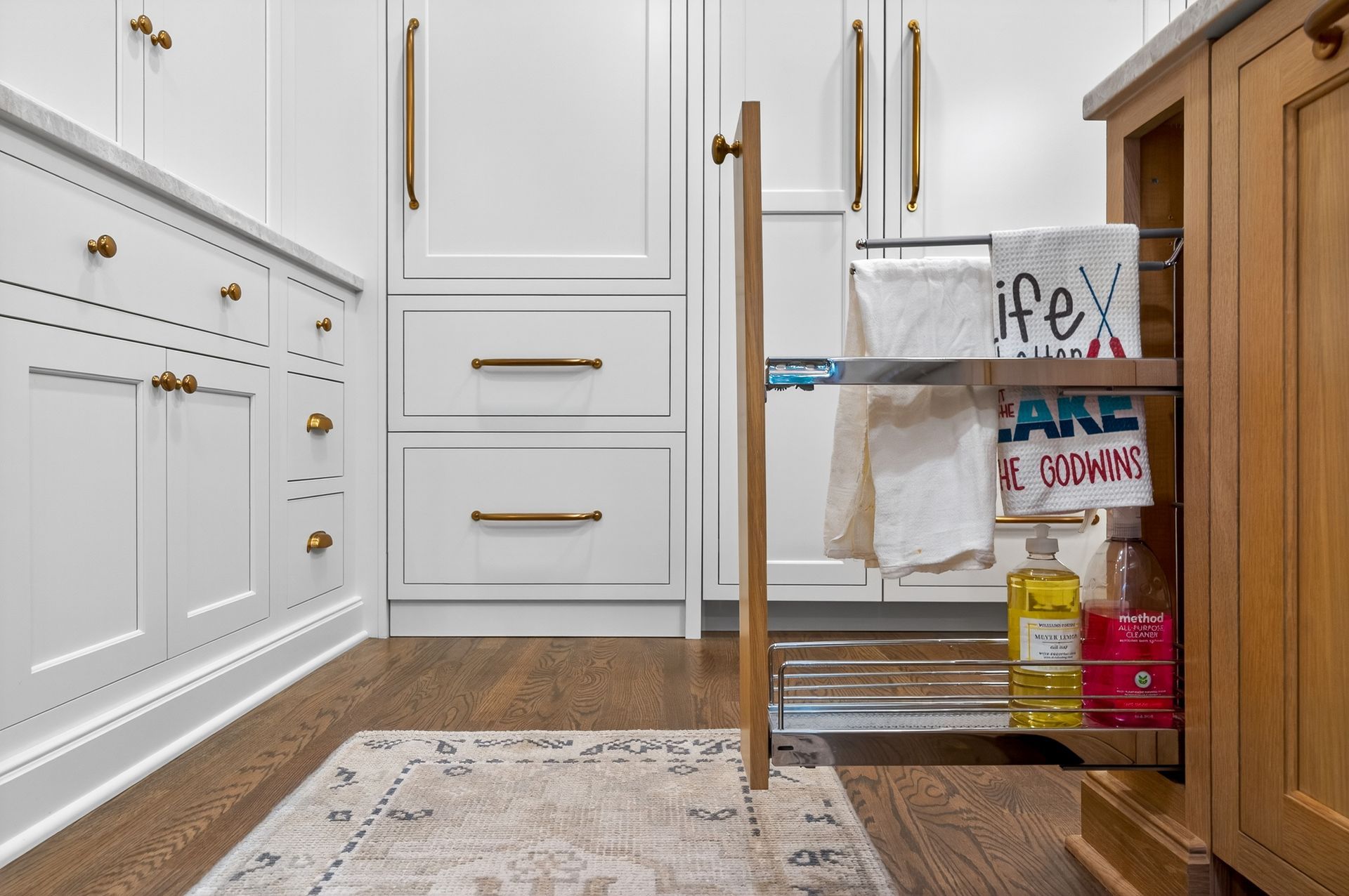 Pull-out cabinet with towels and cleaning supplies next to white cabinets with gold hardware.