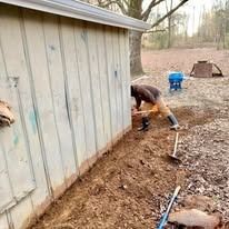 Person digging a trench beside a shed, with pipes and a blue bucket on the dirt ground