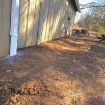 Dirt path beside a beige building with a white downspout, leading to blue bins and trees beyond.