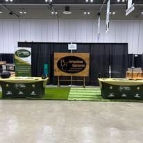 Trade show booth with green table covers, black backdrop, and a central wooden sign under bright lights.