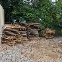 Stacks of wooden pallets piled beside a building in a gravel yard, with trees in the background