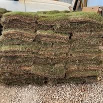Stacked rolls of brown-green sod on gravel, with a vehicle partially visible behind them