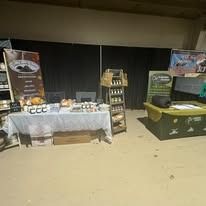 Indoor market booth with packaged goods on white tables, a wooden shelf, and green display banners