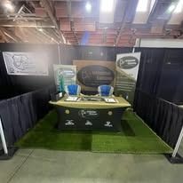 Trade show booth with black drapes, green carpet, and a central table display under overhead lights.