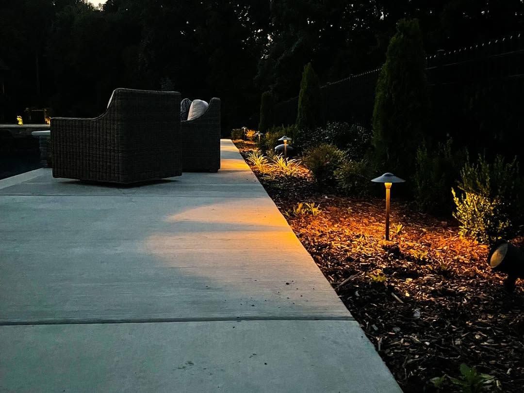 Dimly lit garden walkway beside wicker patio seating, with glowing path lights and shrubs along the edge.
