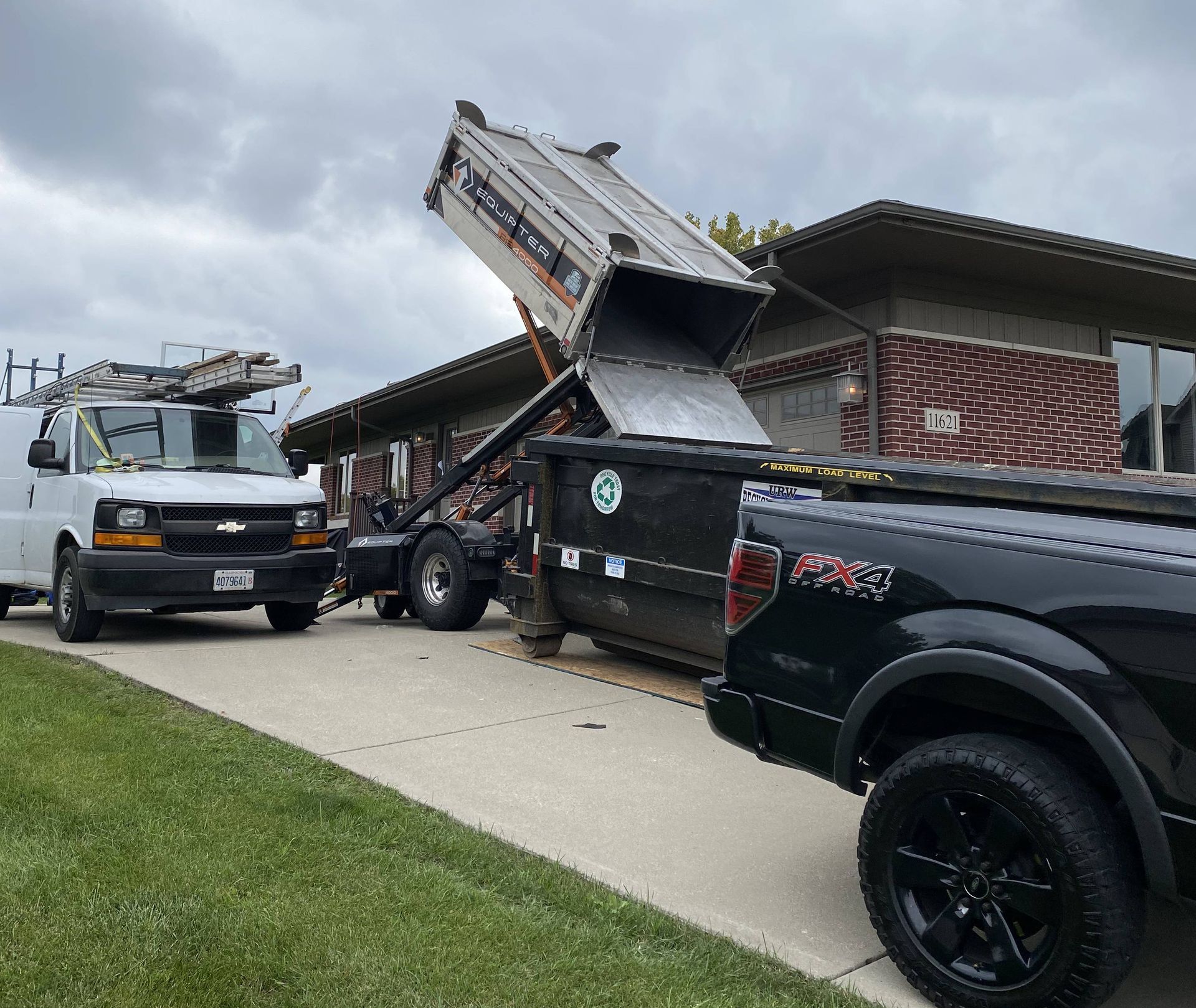 Truck dumping debris into a dumpster next to a house. A work van and pickup truck are also present.
