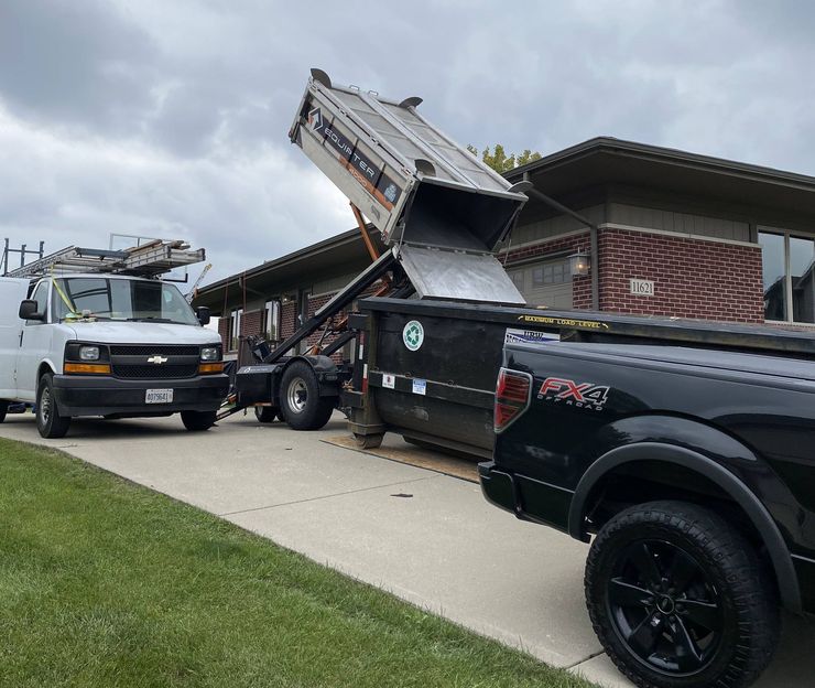 Truck dumping debris into a dumpster next to a house. A work van and pickup truck are also present.