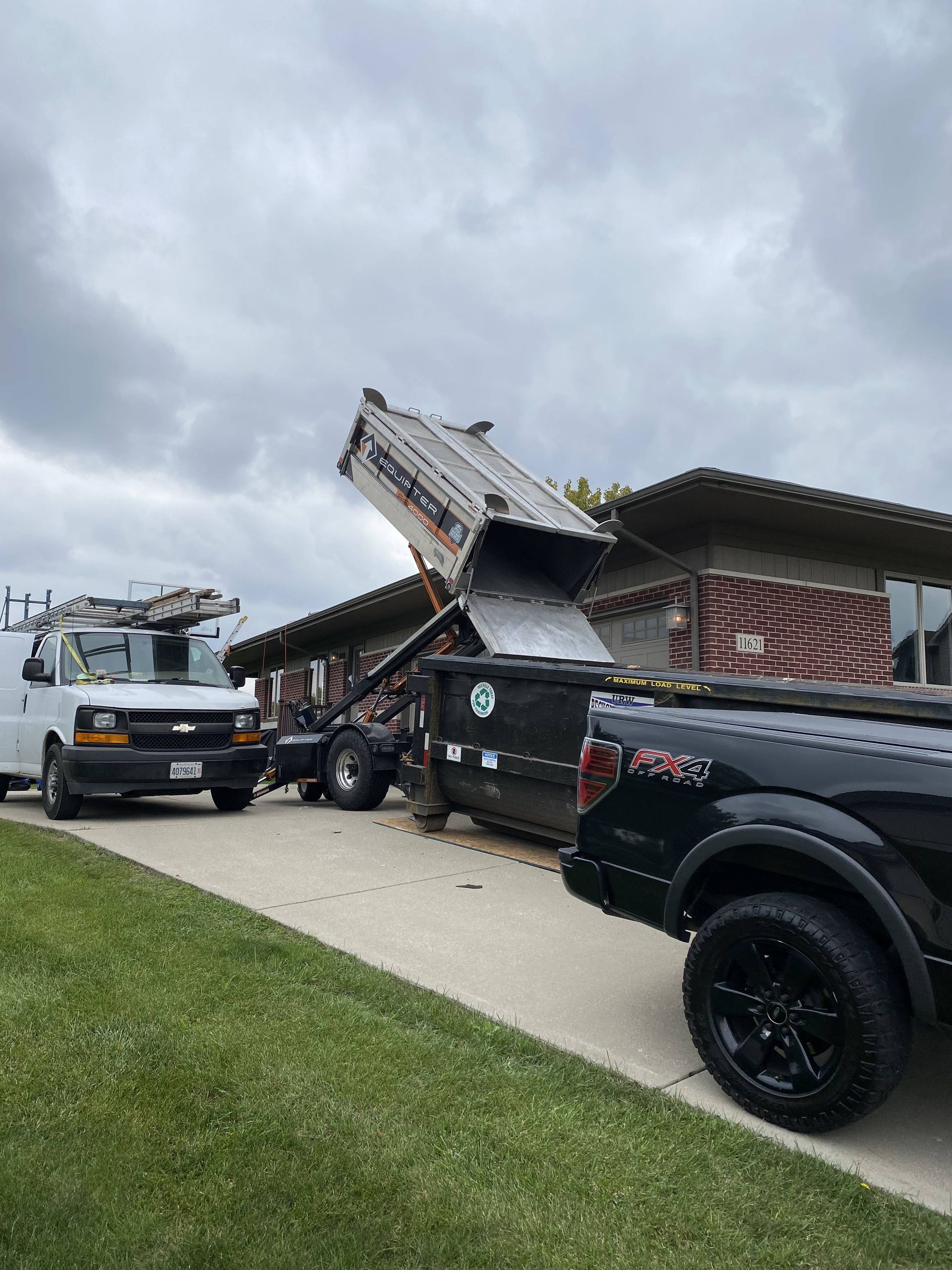 Dump truck emptying debris into a large dumpster in front of a brick house on a cloudy day.