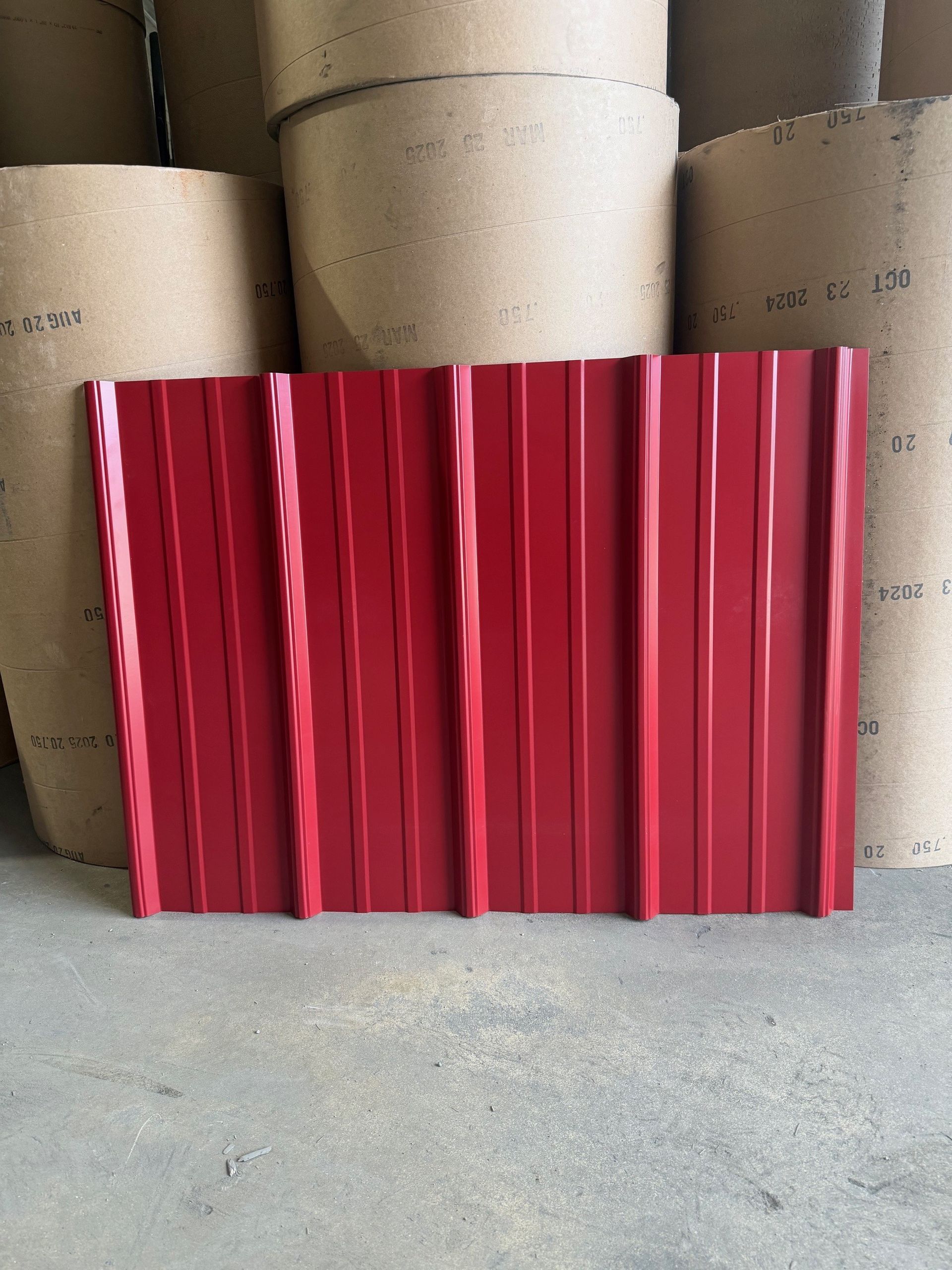 A red, corrugated metal roofing sheet standing upright in front of several large rolls of brown industrial paper.