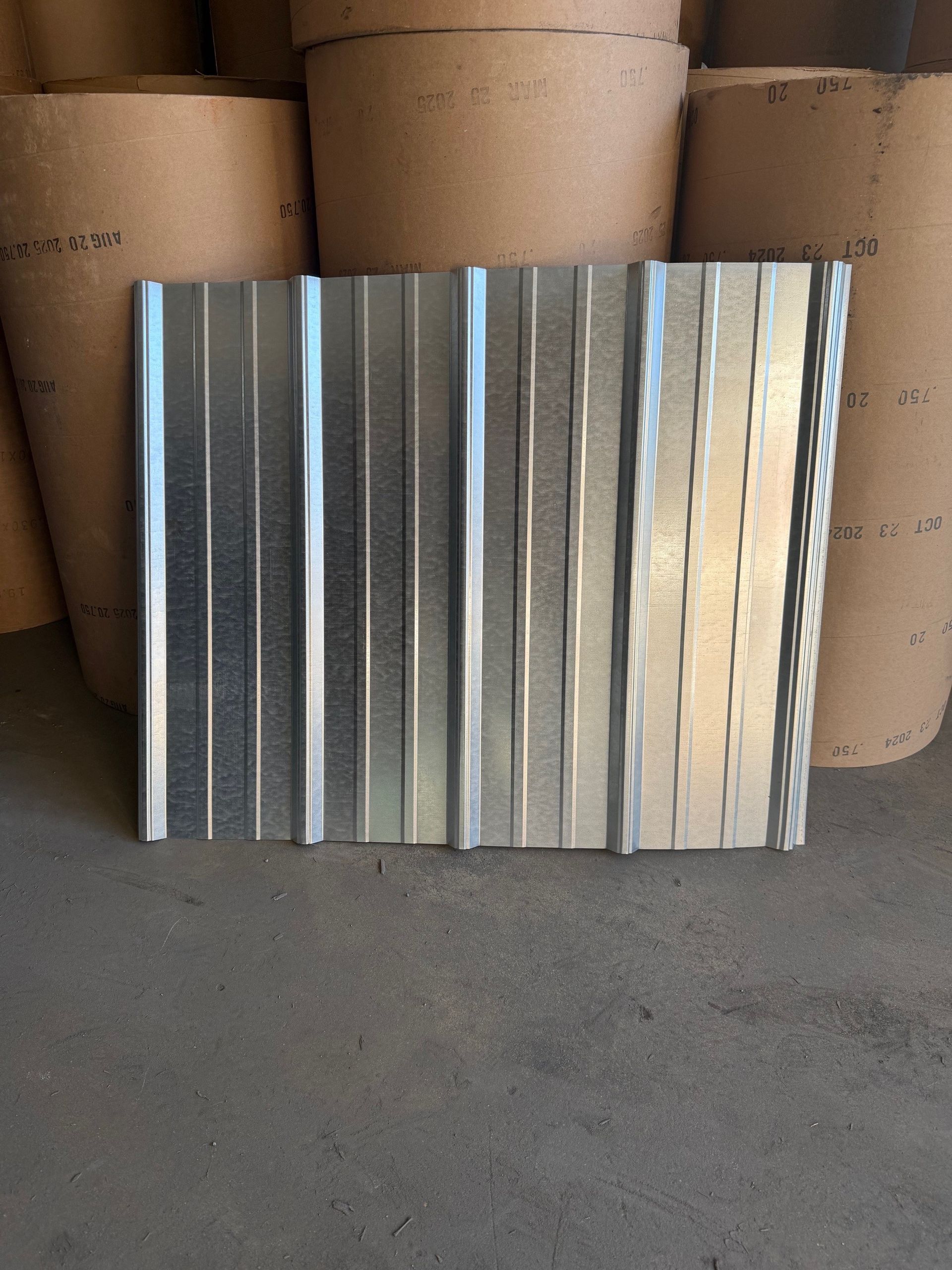 A silver corrugated metal sheet standing vertically in front of several large rolls of brown paper.