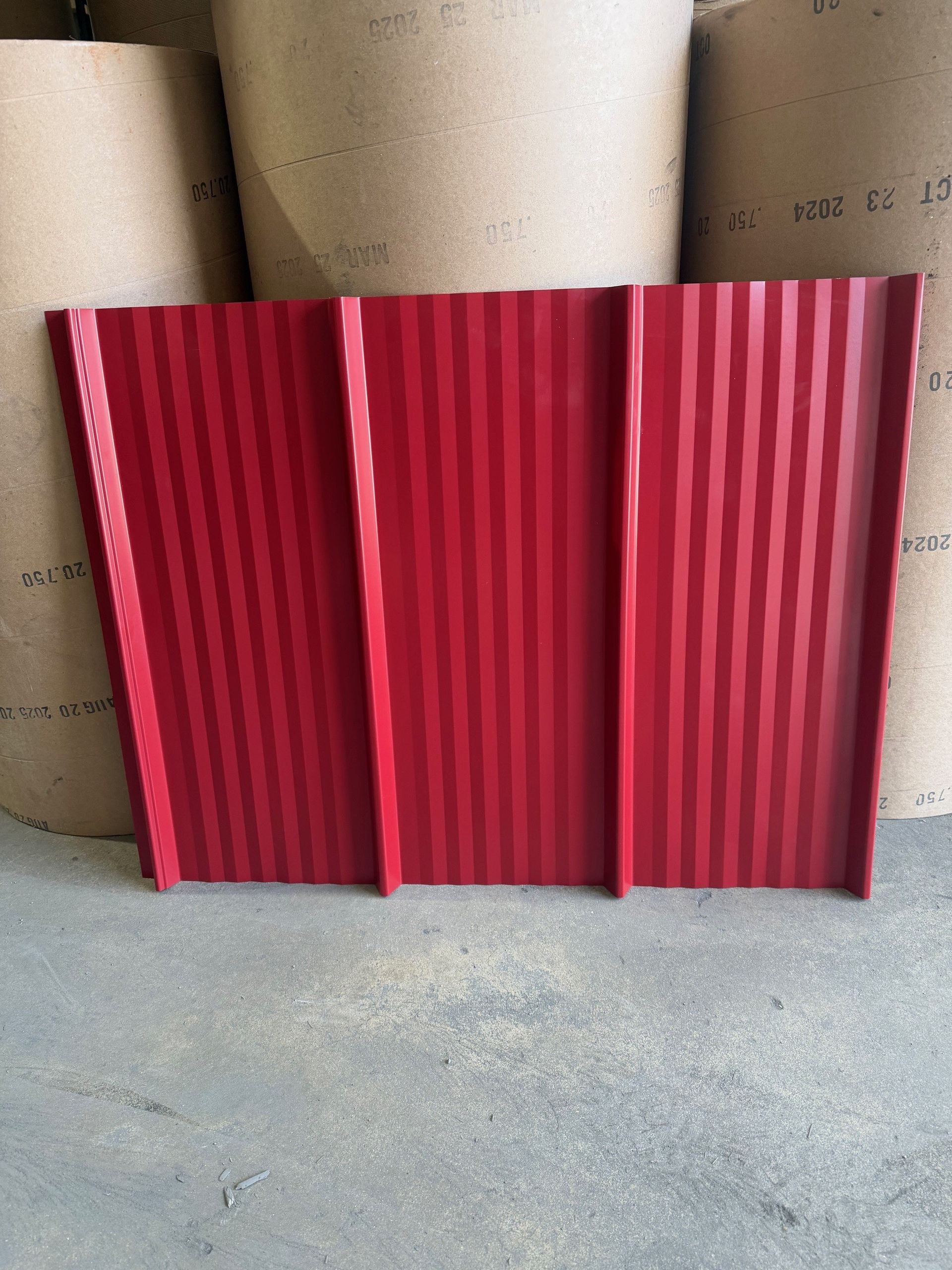 A rectangular, red corrugated metal roofing panel standing upright in front of several large rolls of paper.