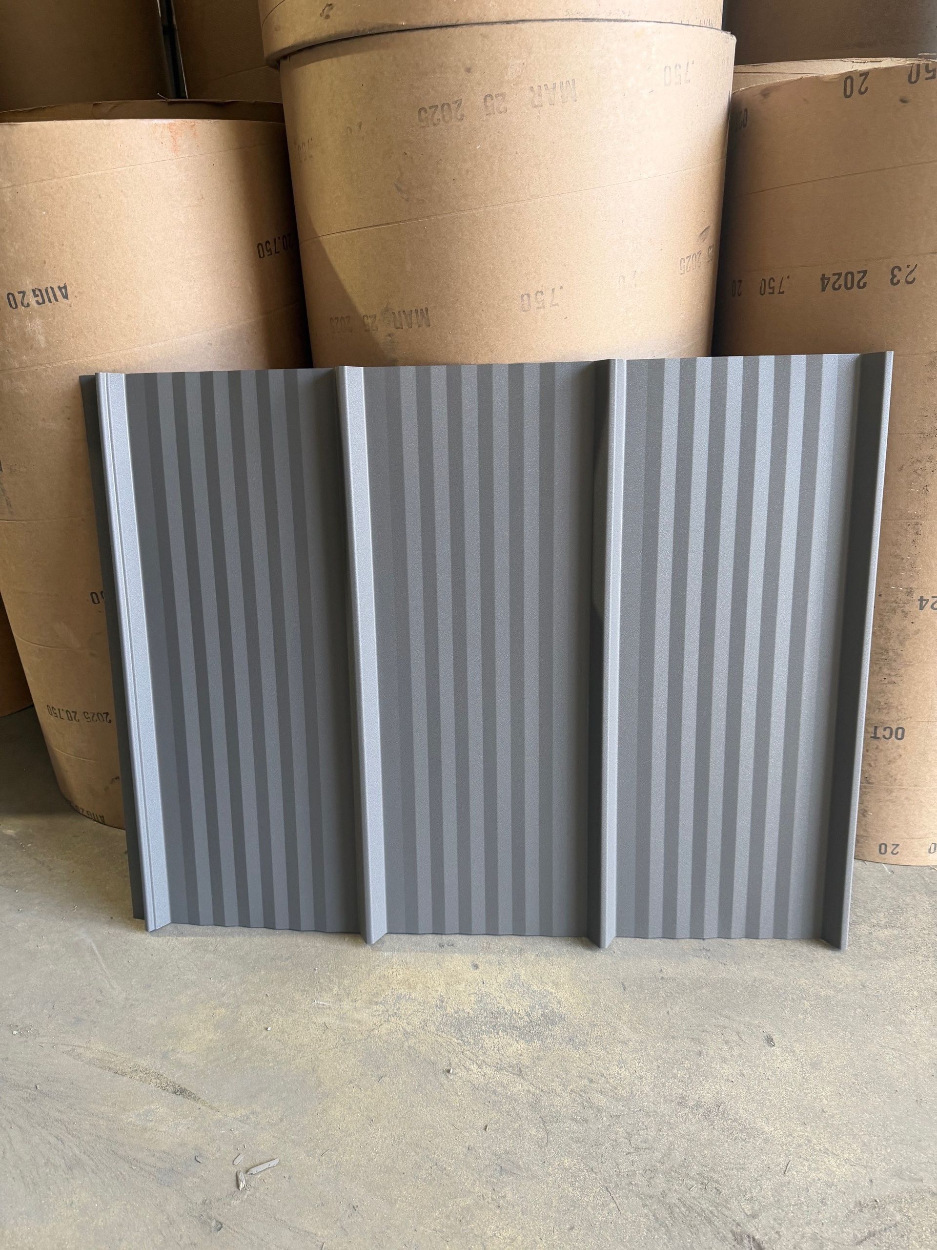 A gray, corrugated metal roofing panel standing upright in front of several large rolls of industrial paper.