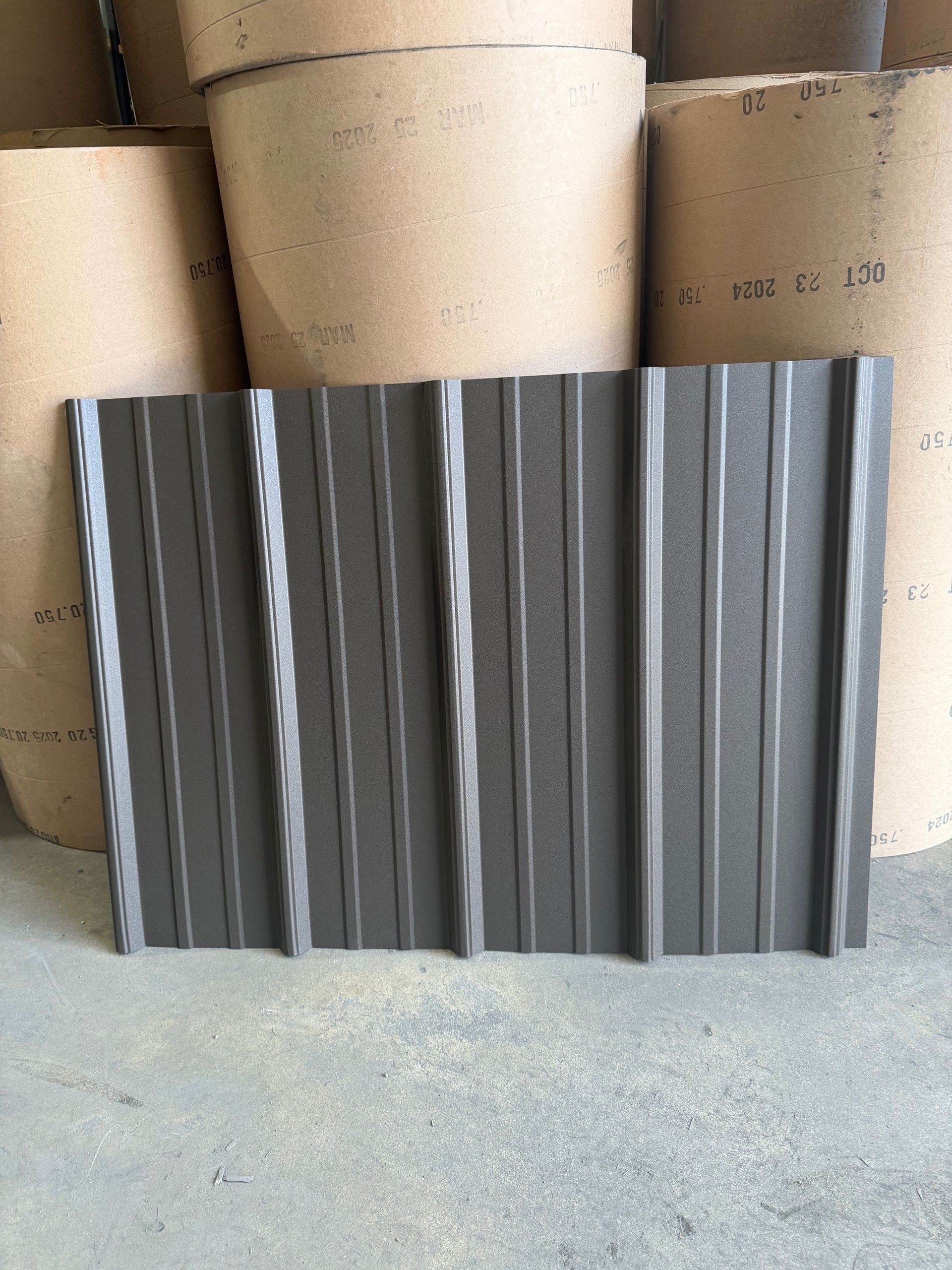 A textured, dark grey metal roofing panel stands upright in front of several large rolls of industrial materials.