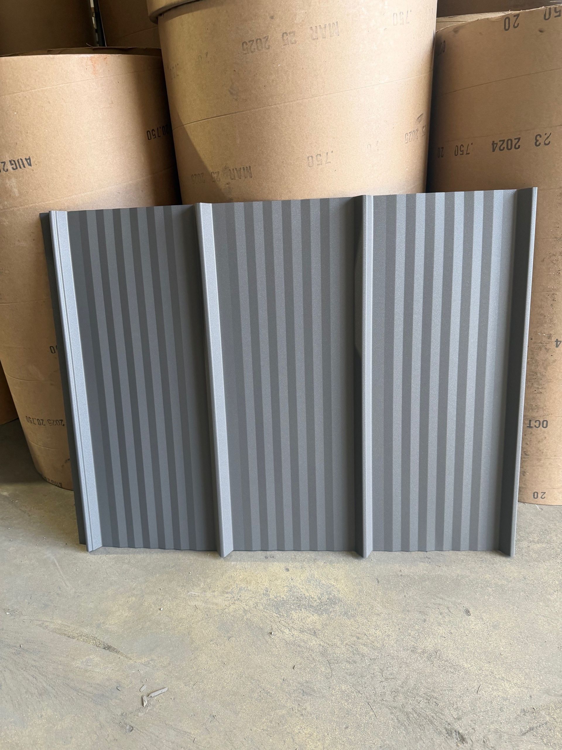 A grey, corrugated metal roofing panel stands vertically in front of large cardboard cylinders in a warehouse setting.