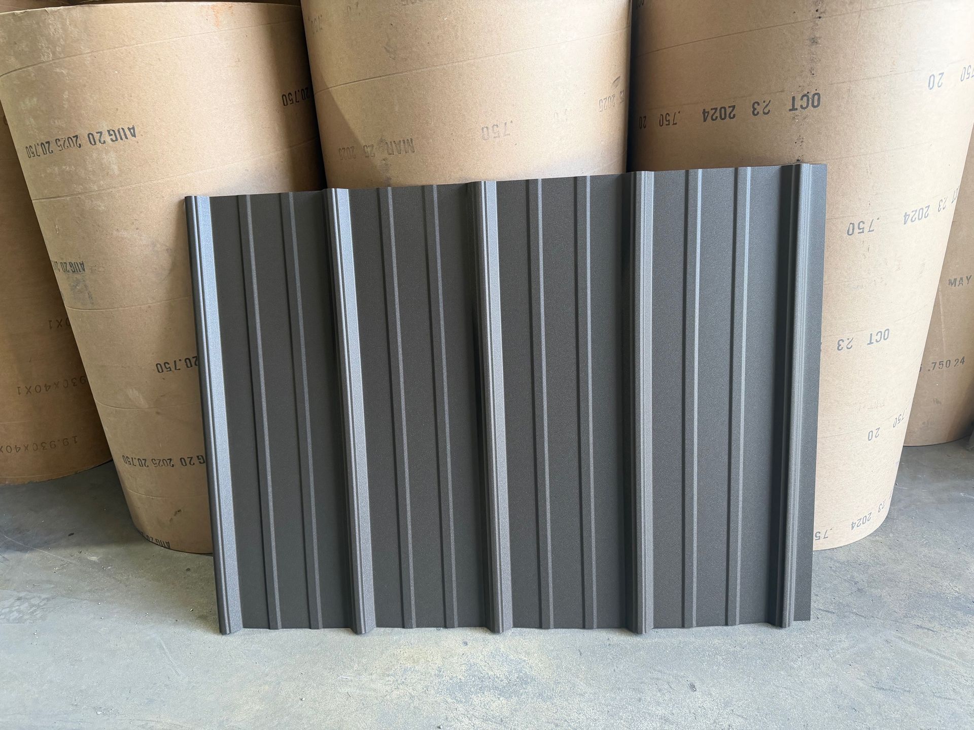 A rectangular sample of dark gray, corrugated metal roofing siding standing against large rolls of craft paper.