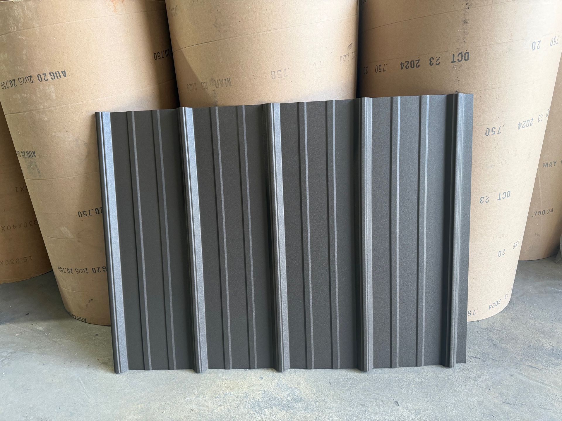 A textured dark gray metal roofing panel standing upright in front of several large rolls of paper material.