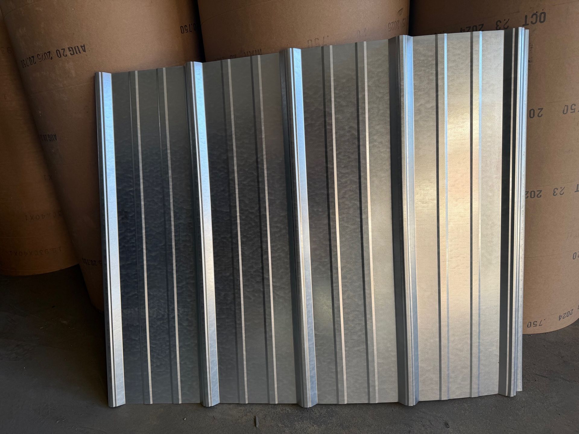 A rectangular galvanized steel roofing panel with vertical ribbing, shown against a background of rolled industrial paper.