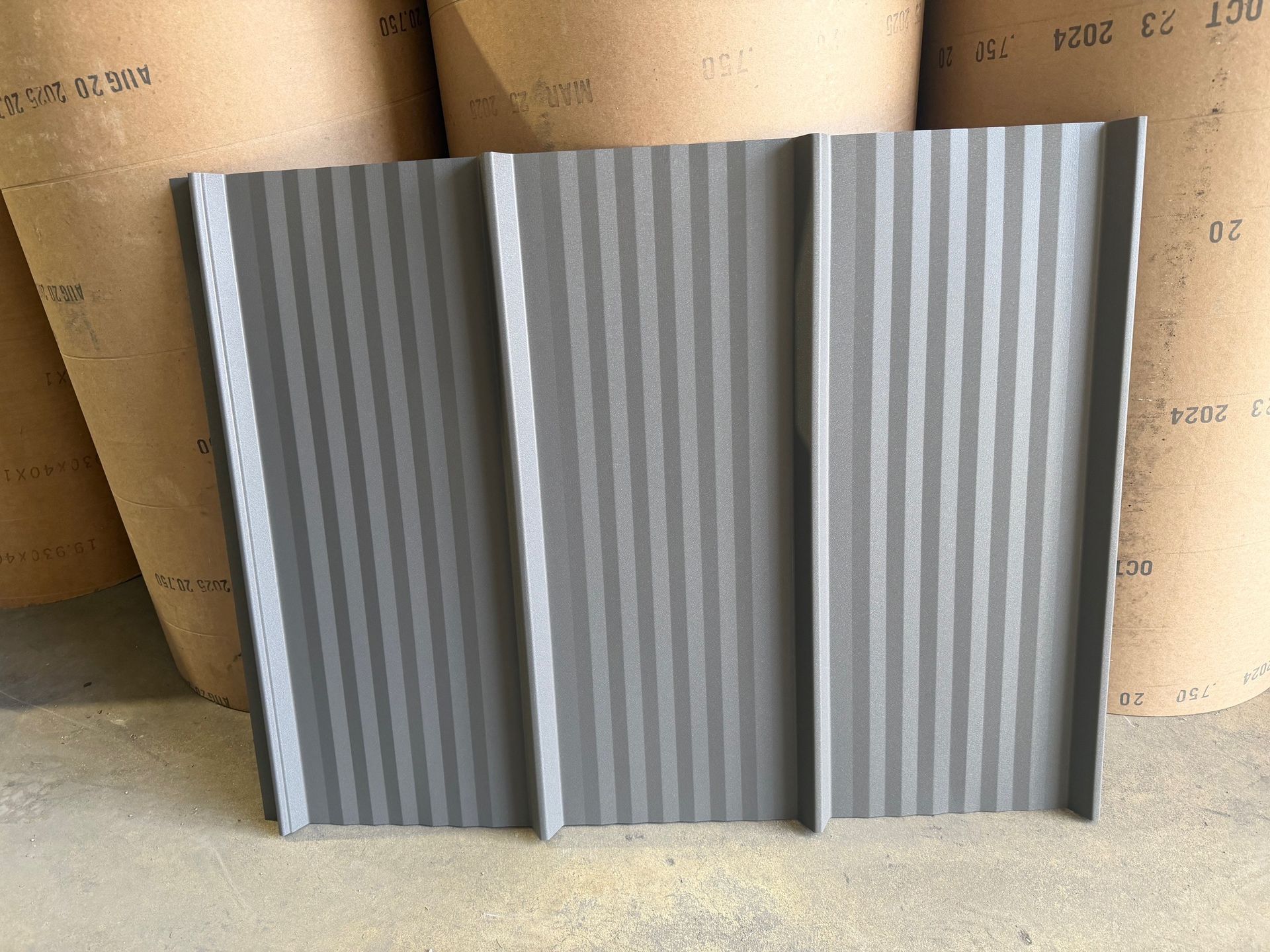 A grey, metal roofing panel with vertical raised ribs, standing in front of large rolls of industrial paper.