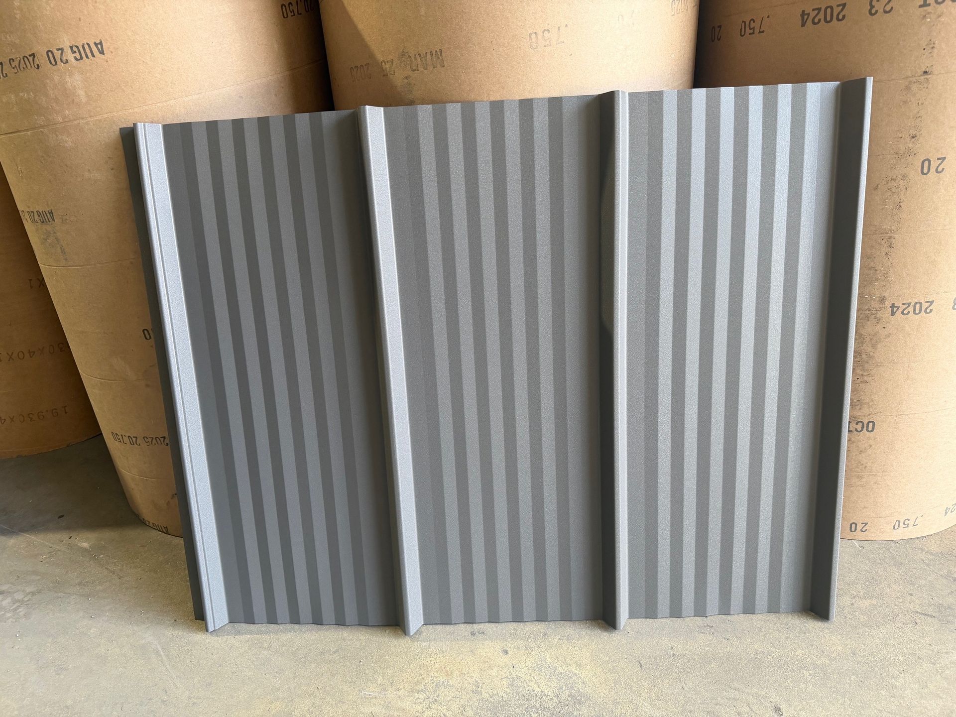 A gray, metal roofing panel with a ribbed, corrugated texture, standing upright in front of several large paper rolls.