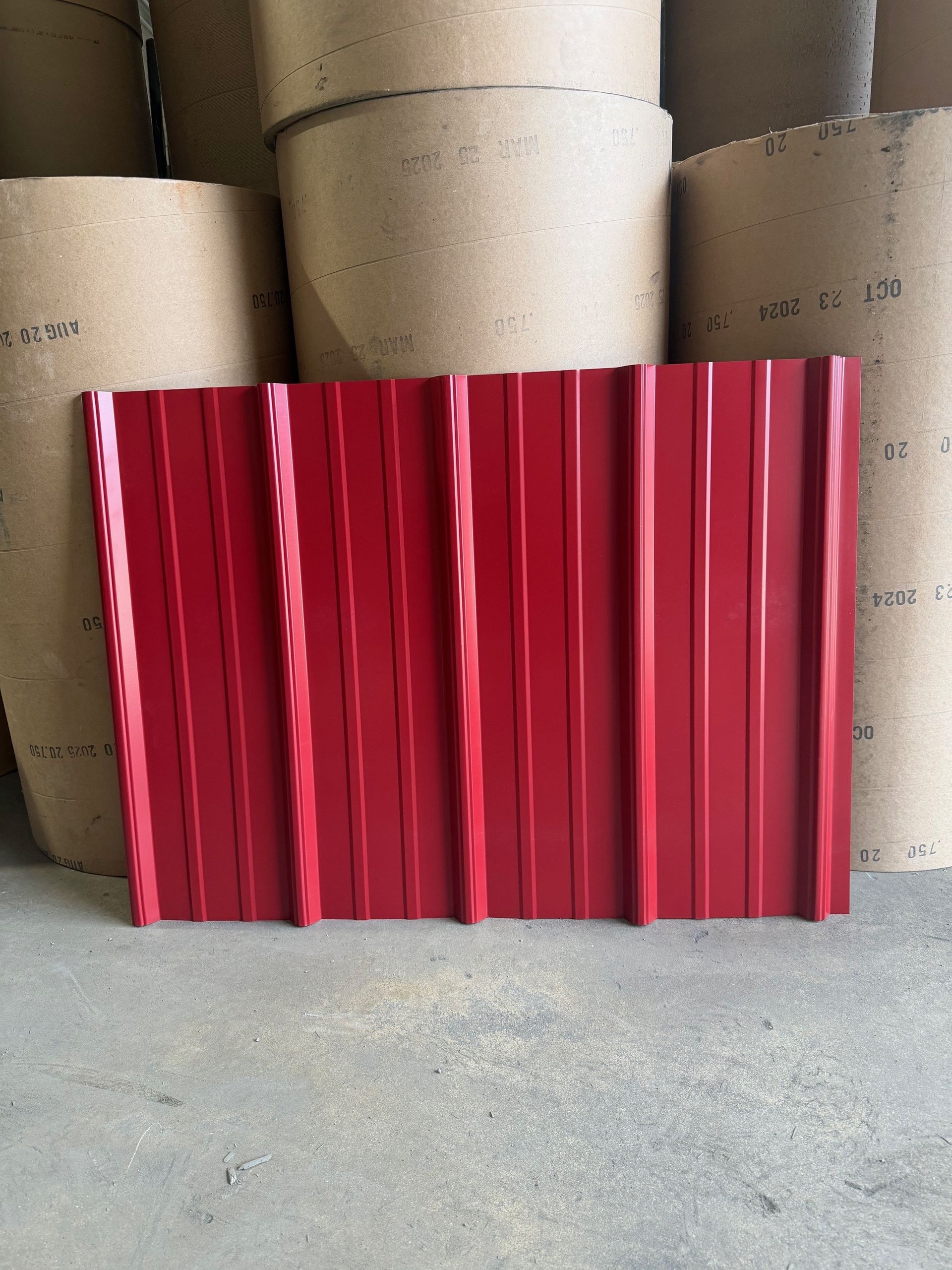 A rectangular piece of deep red corrugated metal roofing panel standing in front of several large rolls of paper.