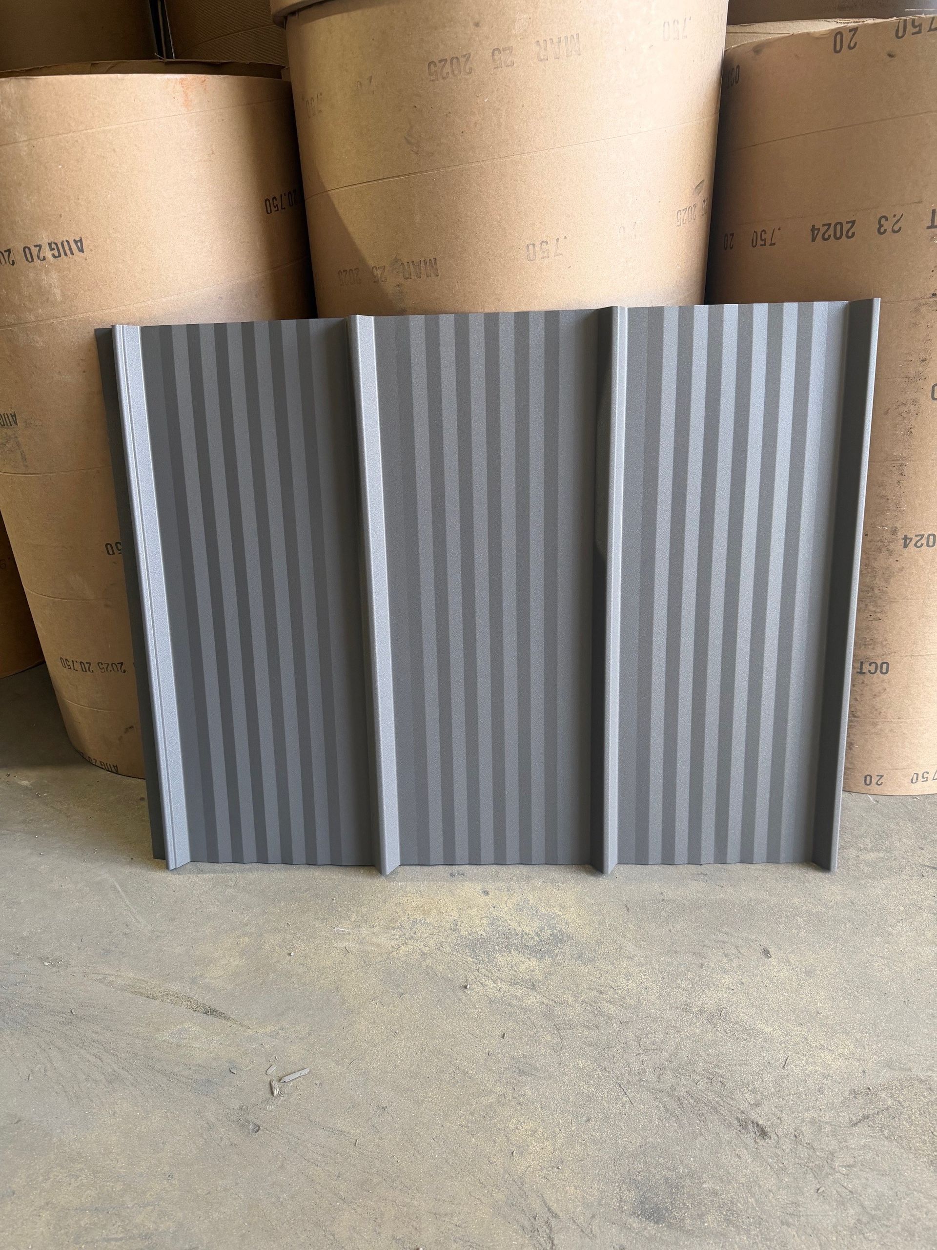 A dark grey, corrugated metal roofing panel stands upright in front of several large paper rolls in a warehouse.