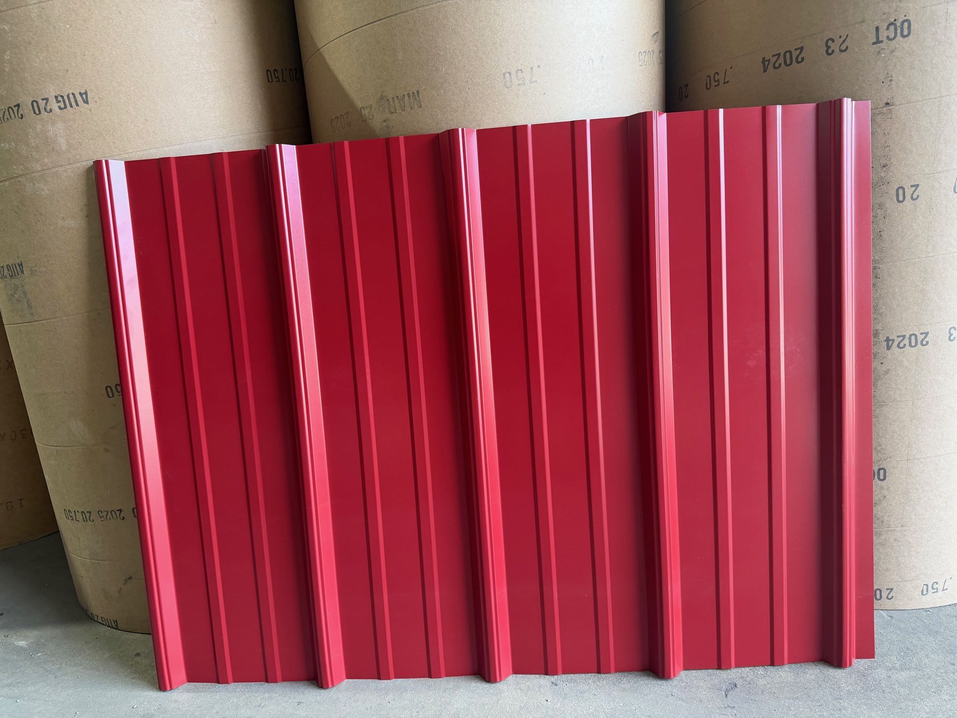 A single panel of bright red, corrugated metal roofing with vertical ribs standing in front of large industrial rolls.