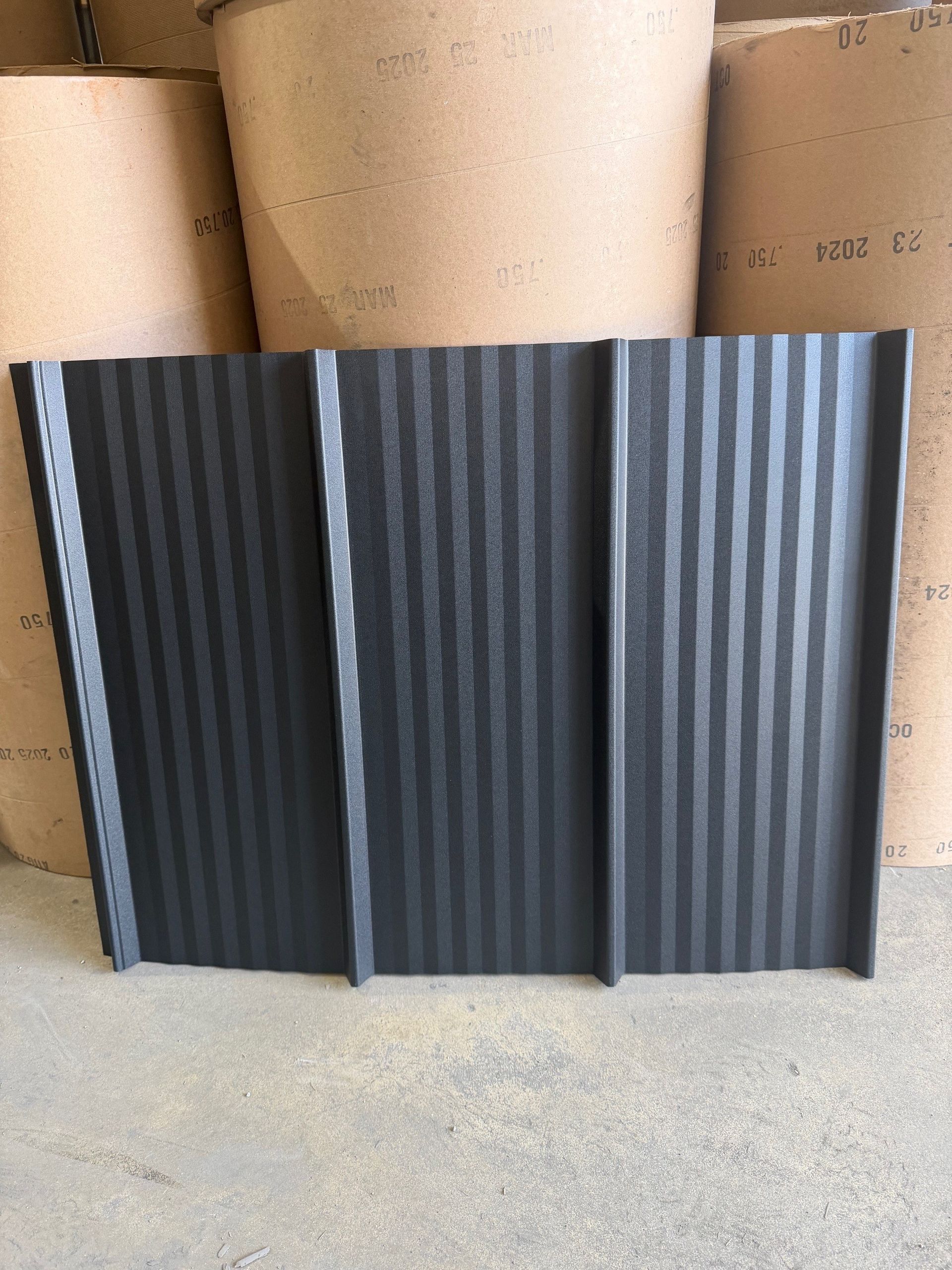 A dark gray, corrugated metal roofing panel standing vertically against a background of large, tan industrial rolls.