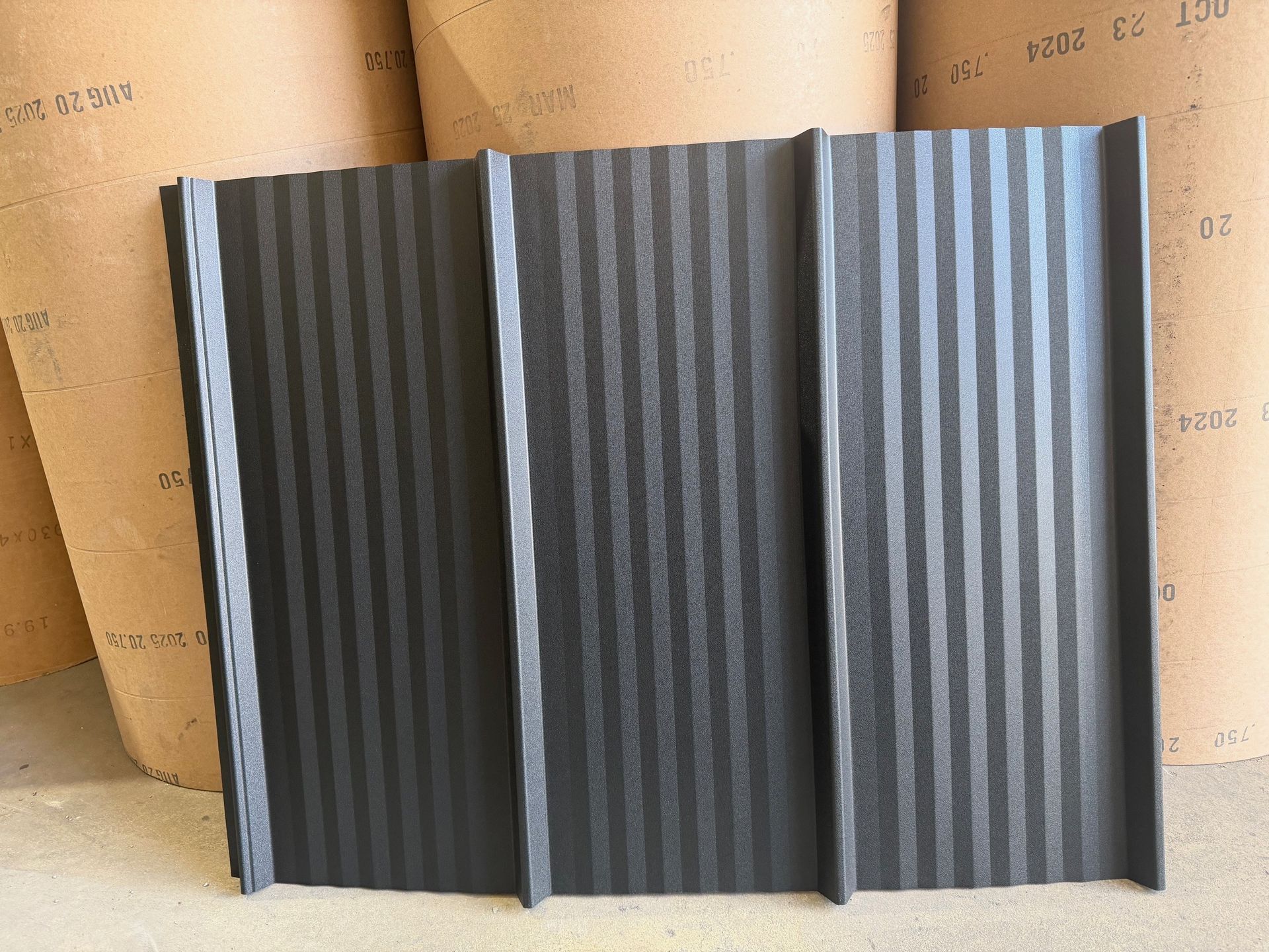 A rectangular, charcoal-colored metal roof panel with vertical ridges standing in front of large paper rolls.