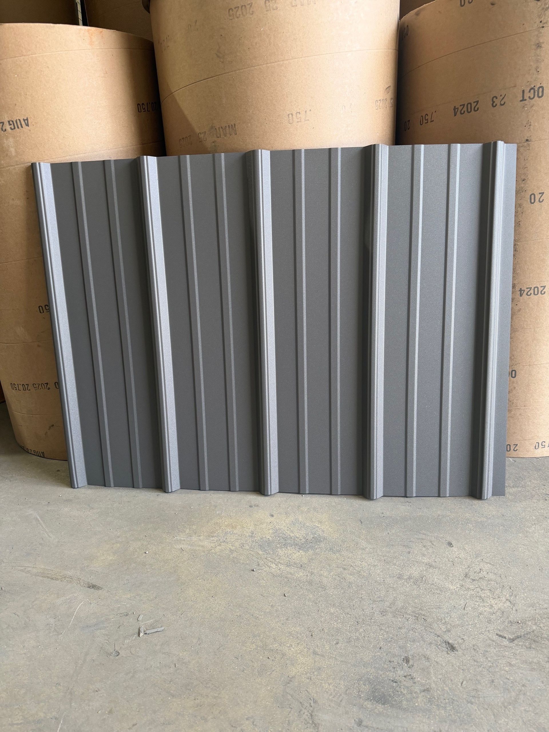 A gray, corrugated metal roofing panel stands vertically in front of large rolls of industrial paper in a warehouse.