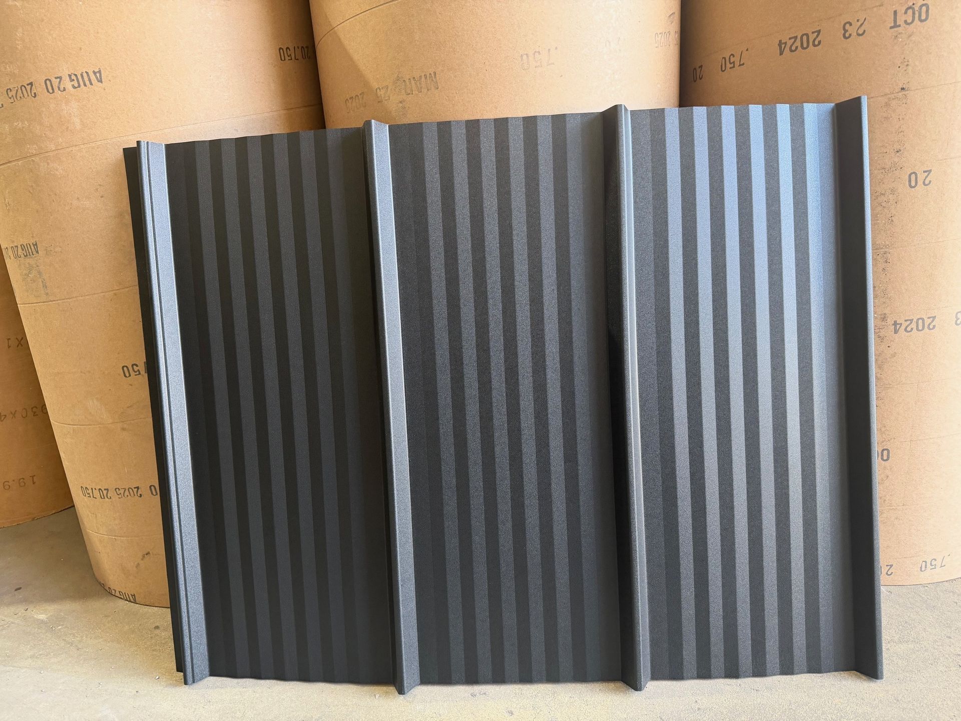A rectangular, charcoal-grey metal roofing panel with vertical corrugated ribs, positioned against large paper rolls.
