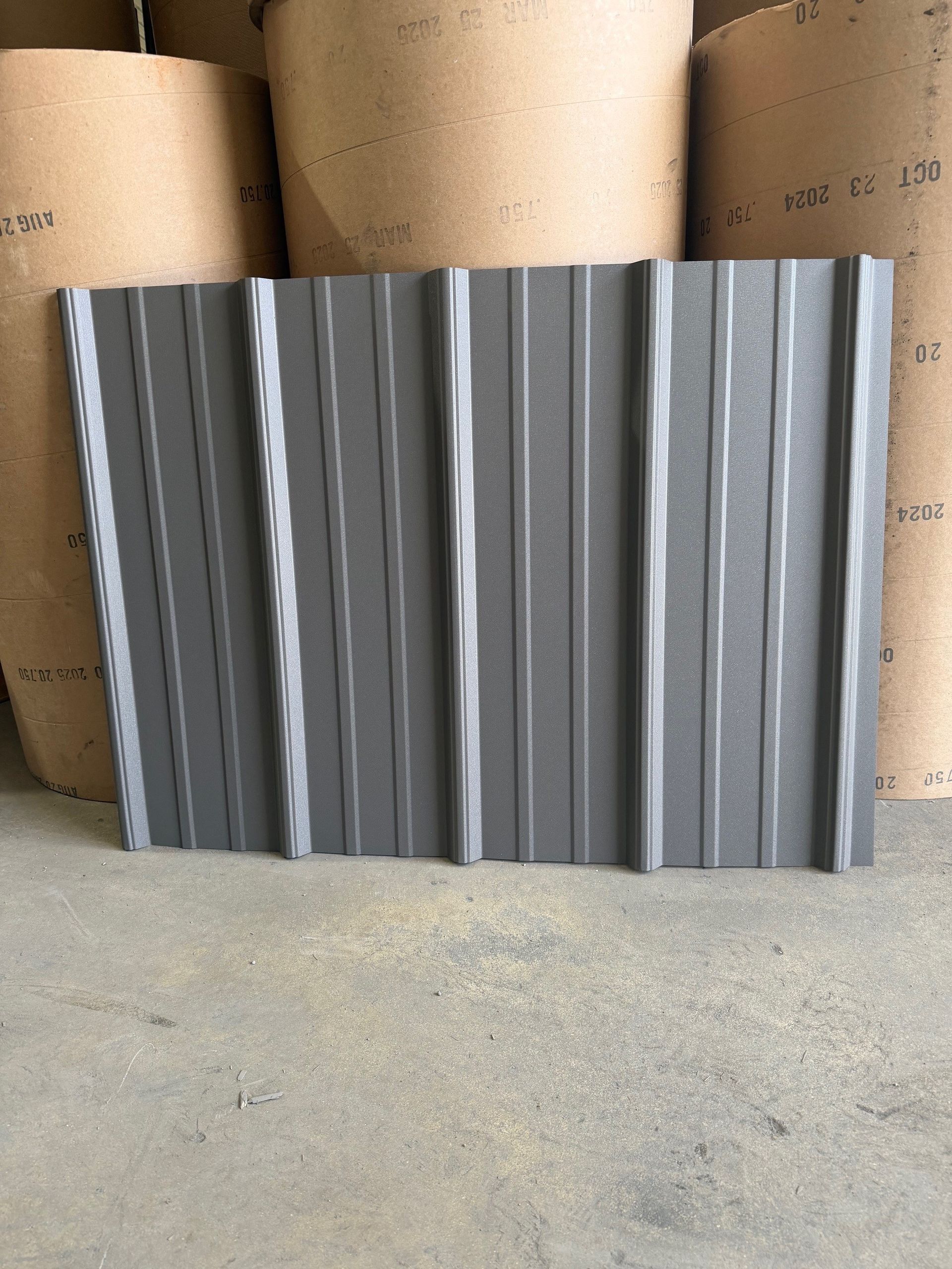 A dark grey corrugated metal roofing panel stands vertically against a backdrop of several large, stacked paper rolls.