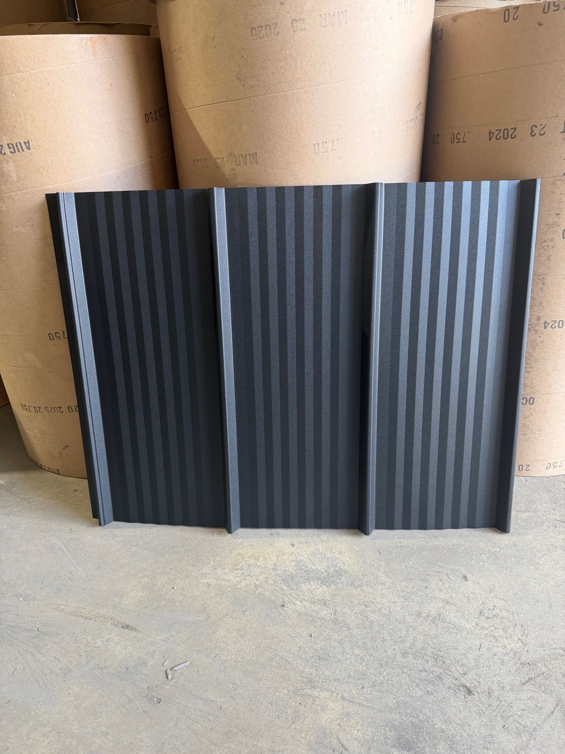 A black, textured, rectangular metal roofing panel with three vertical ribs, standing against large rolls of paper.