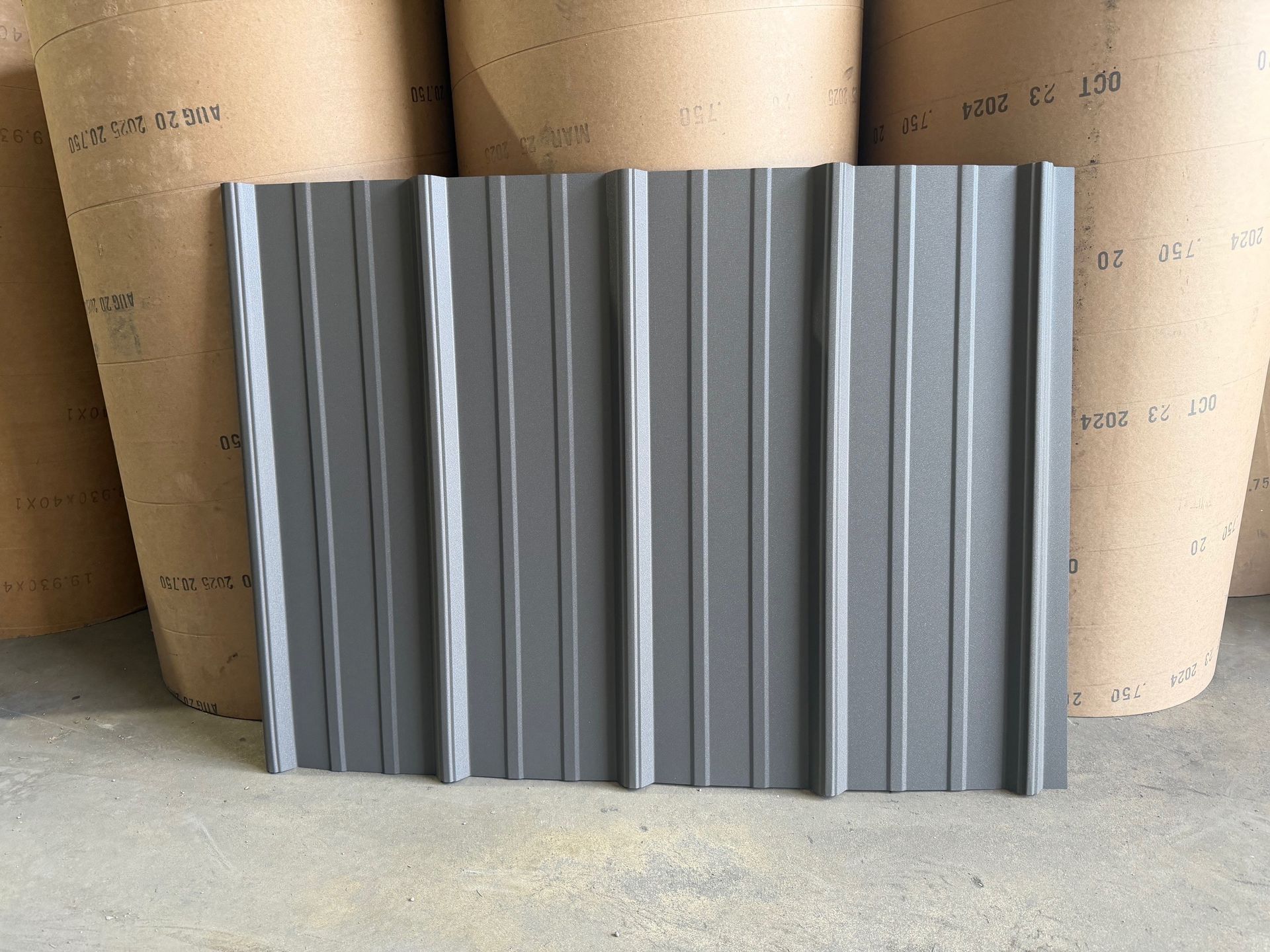 A rectangular panel of dark gray, vertically ribbed metal roofing standing upright against large rolls of paper.