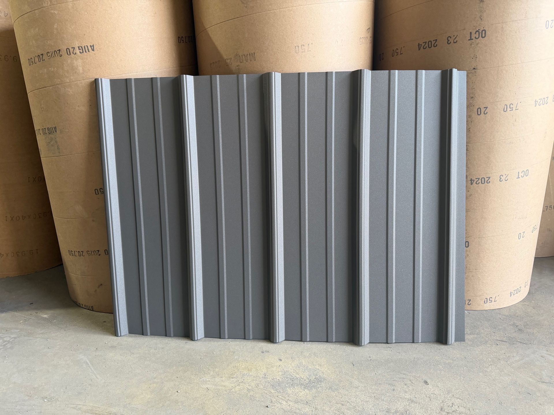 A dark gray metal roofing panel with vertical ribbing, standing upright in front of large rolls of industrial paper.