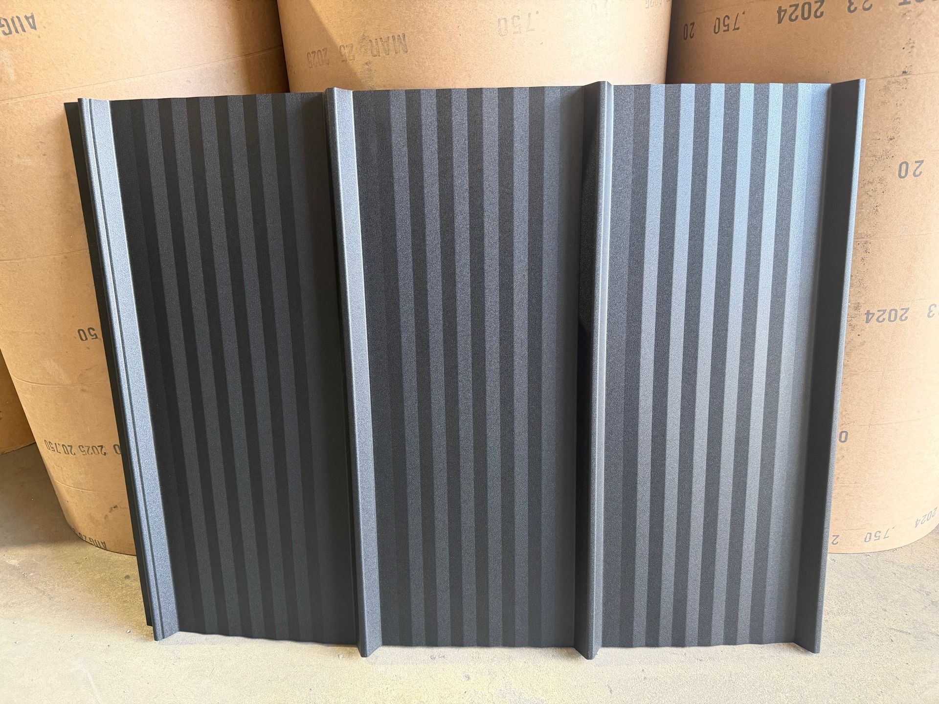 A dark gray, textured metal panel with three vertical raised ribs, standing upright against a light brown backdrop.