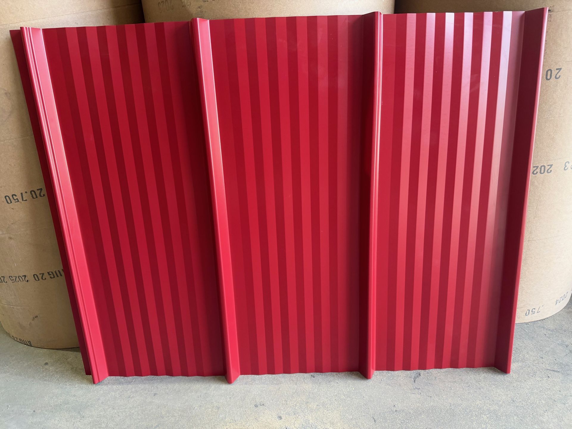 A vertical red metal roofing panel with three prominent raised ridges, standing against a background of paper rolls.
